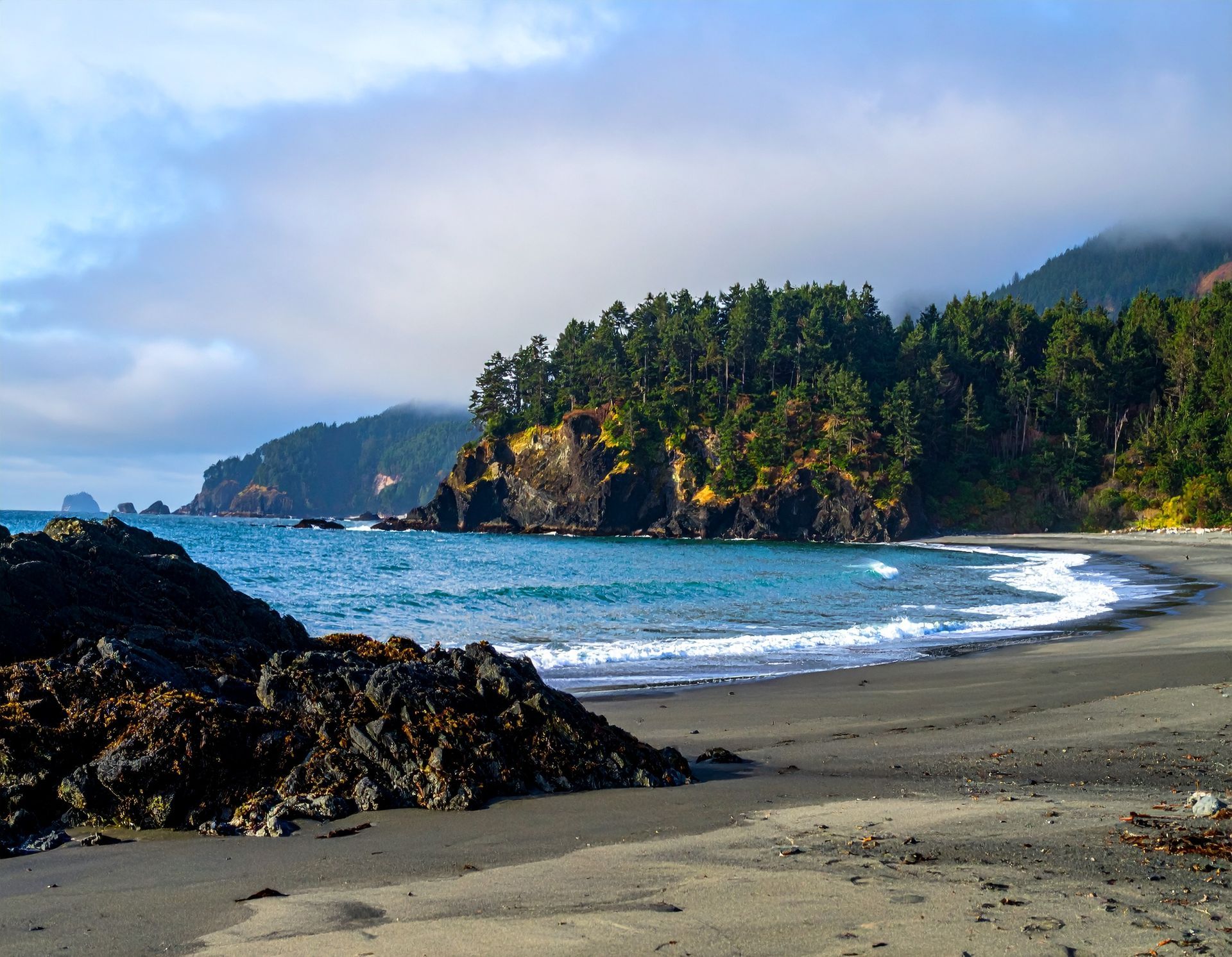 Rocky tropical beach with waves, sandy shore, cliffs, and dense green trees under a cloudy blue sky