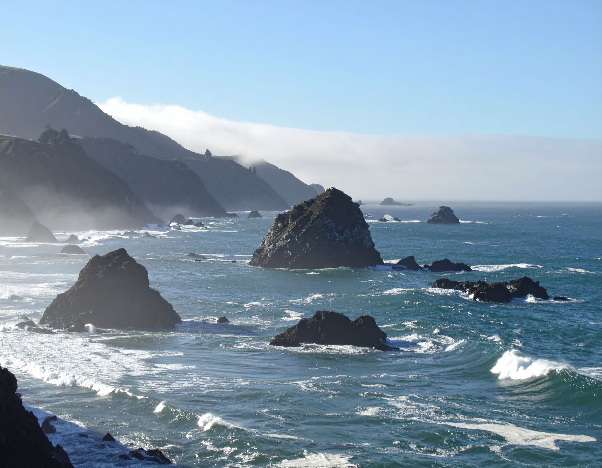 Rocky sea coast with waves, offshore sea stacks, and hazy cliffs under a blue sky