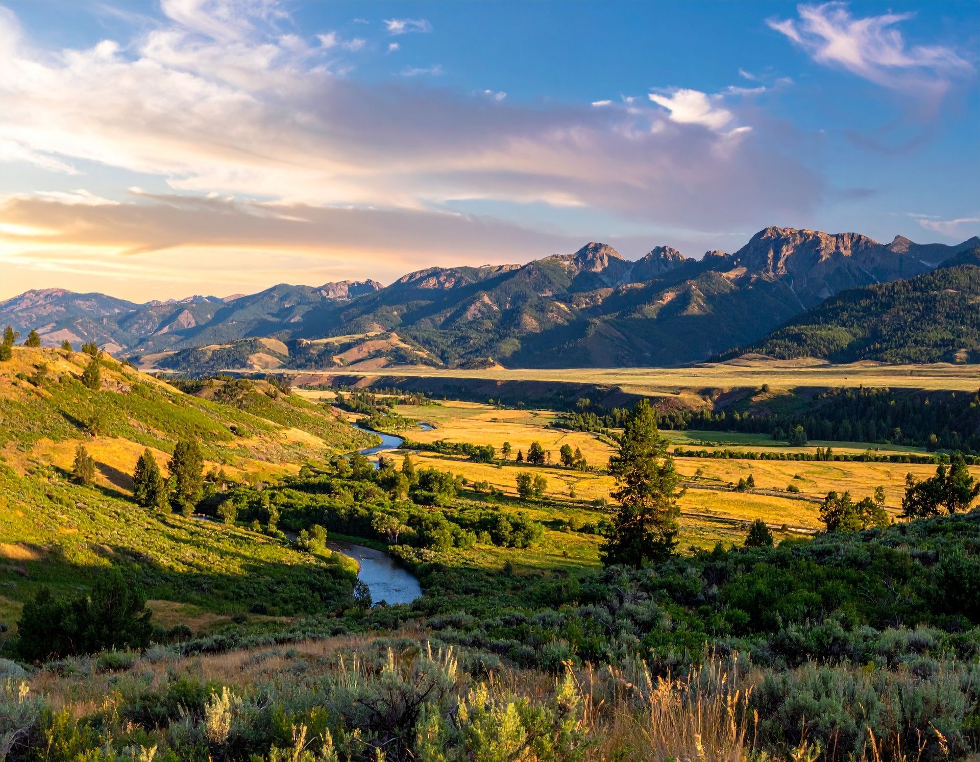 Sunlit mountain valley with a winding stream, golden fields, and rugged peaks under a pink-blue sky