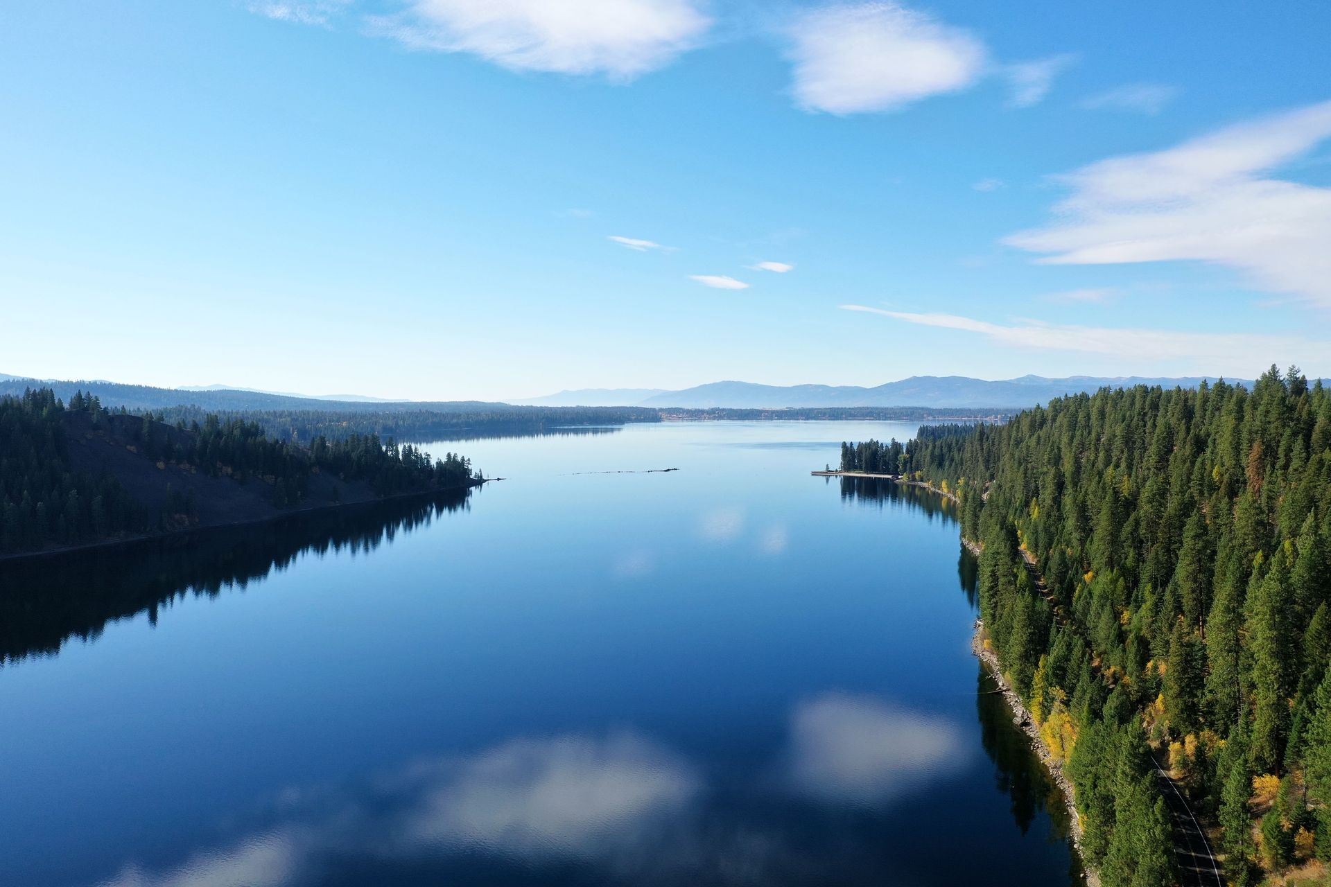 A calm blue lake bordered by forested shores under a bright sky with distant mountains