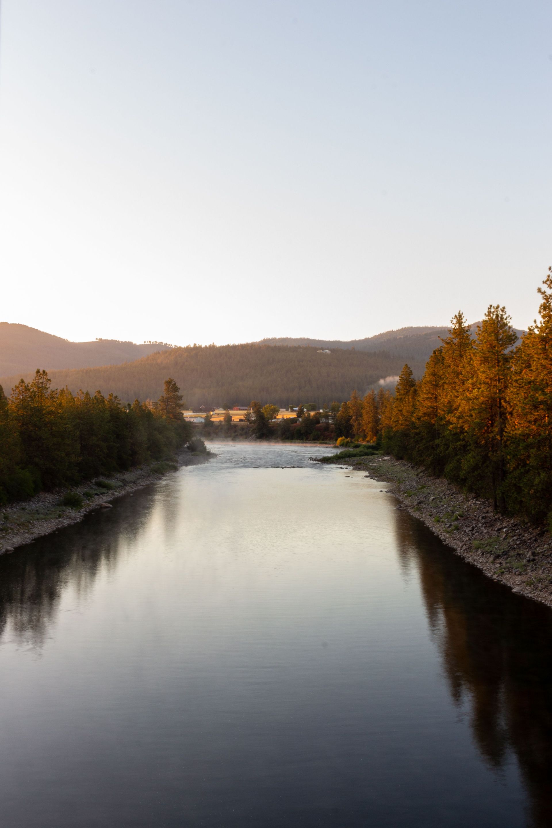 Calm river at sunset with tree-lined banks and distant hills under a clear sky
