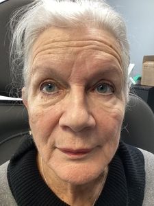 Close-up portrait of a woman with gray hair and a neutral expression, indoors against a dark chair.