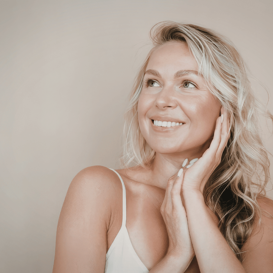 Smiling blonde woman in a white top, looking up with hands on her cheek against a beige background