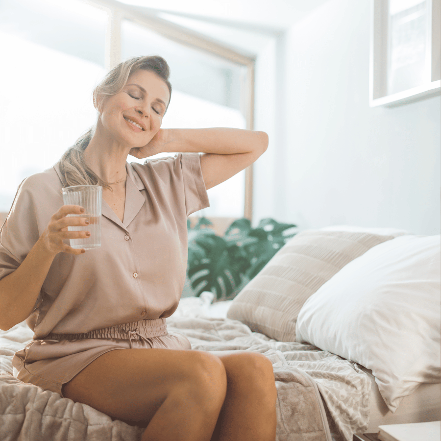 Smiling woman on a bed massaging her neck while holding a glass of water in a bright bedroom
