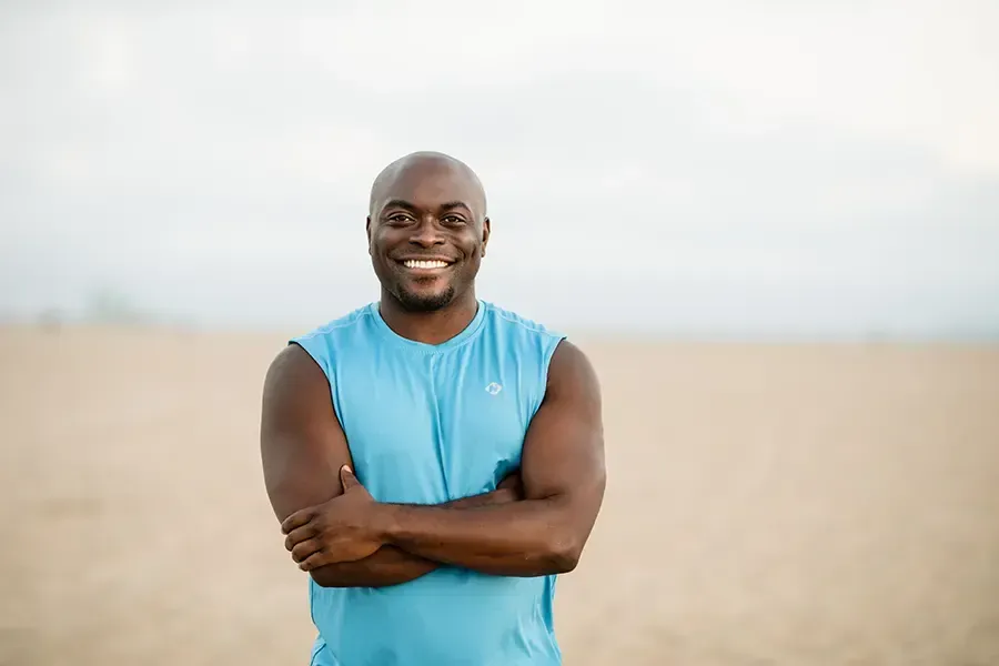 Smiling man in a blue sleeveless shirt standing with arms crossed outdoors in a field