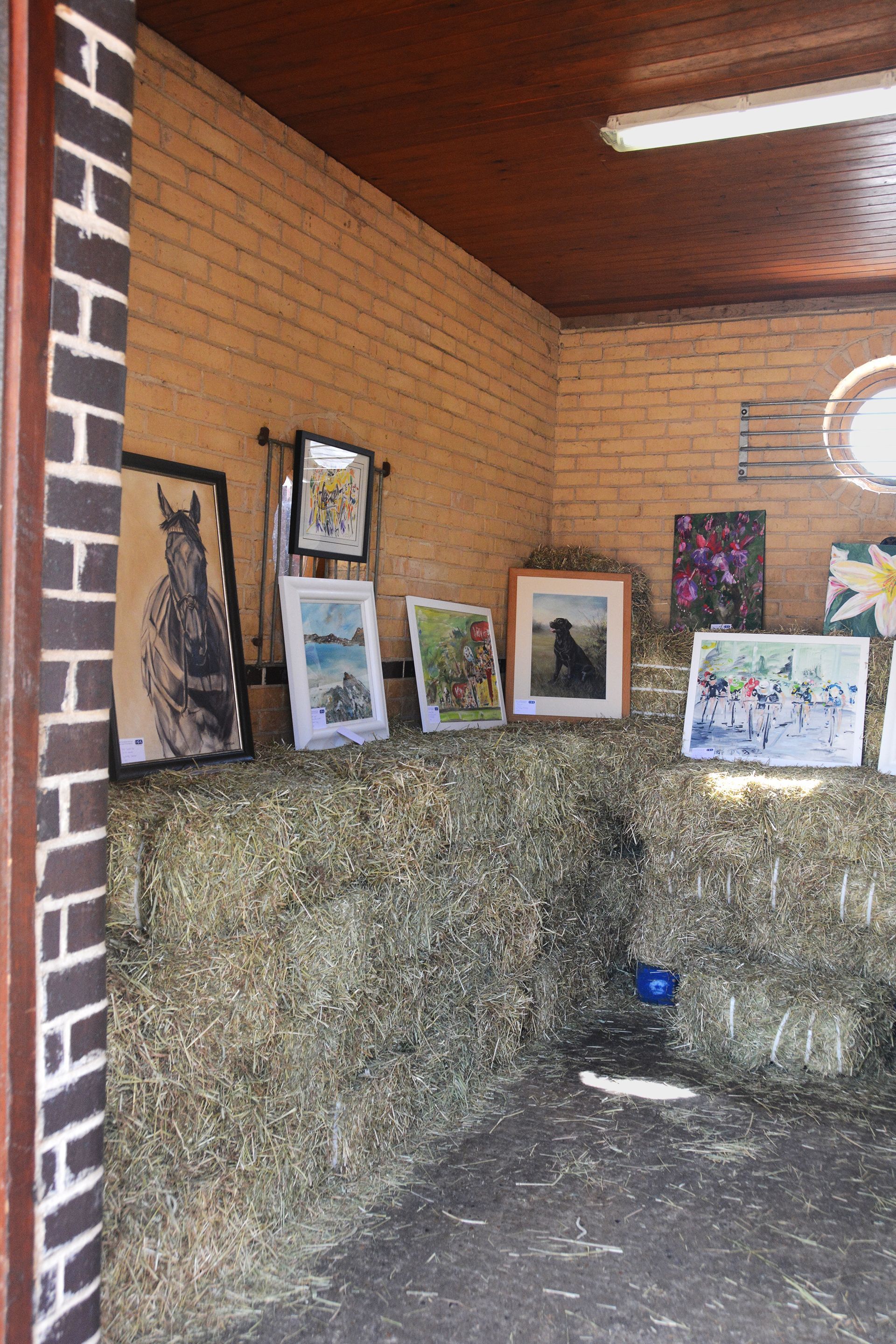 Artwork displayed on hay bales inside a brick-walled stable.