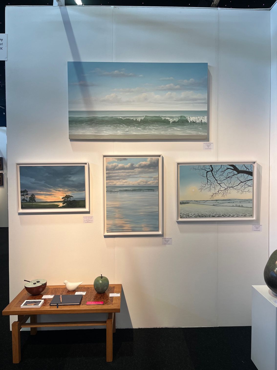 Art gallery display with four seascape paintings. A small wooden table with decor sits in front.