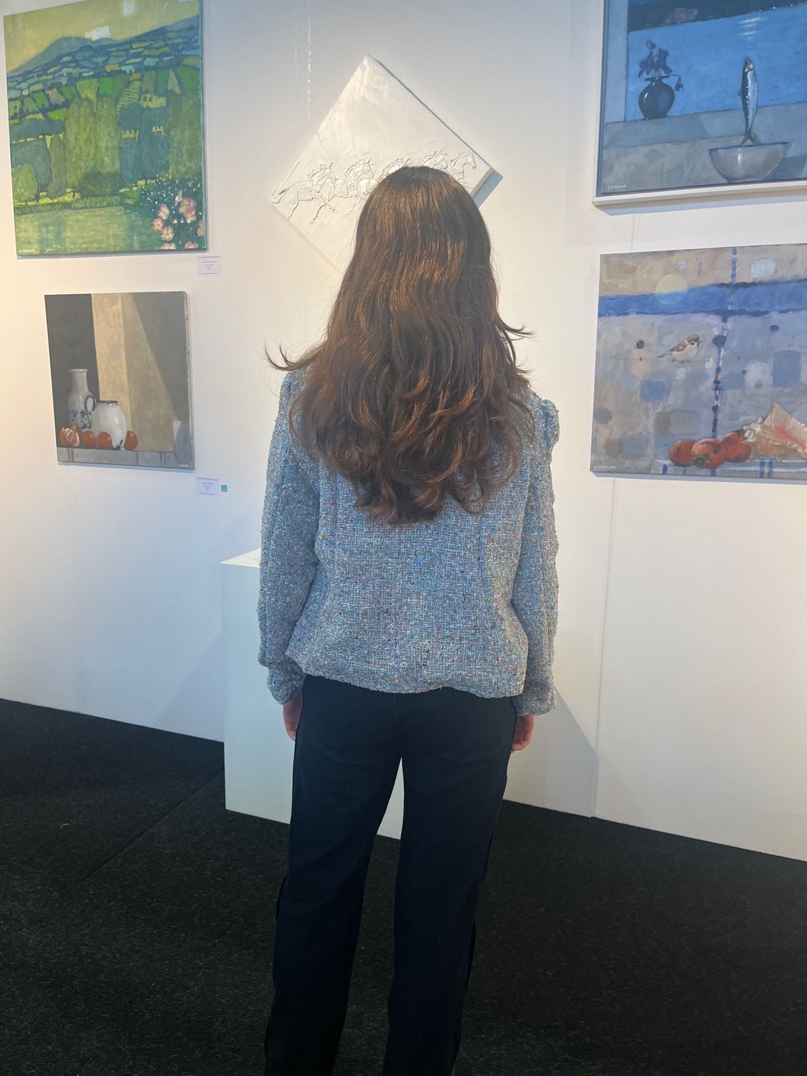 Woman in gallery, looking at paintings on wall. She wears a gray sweater and black pants.