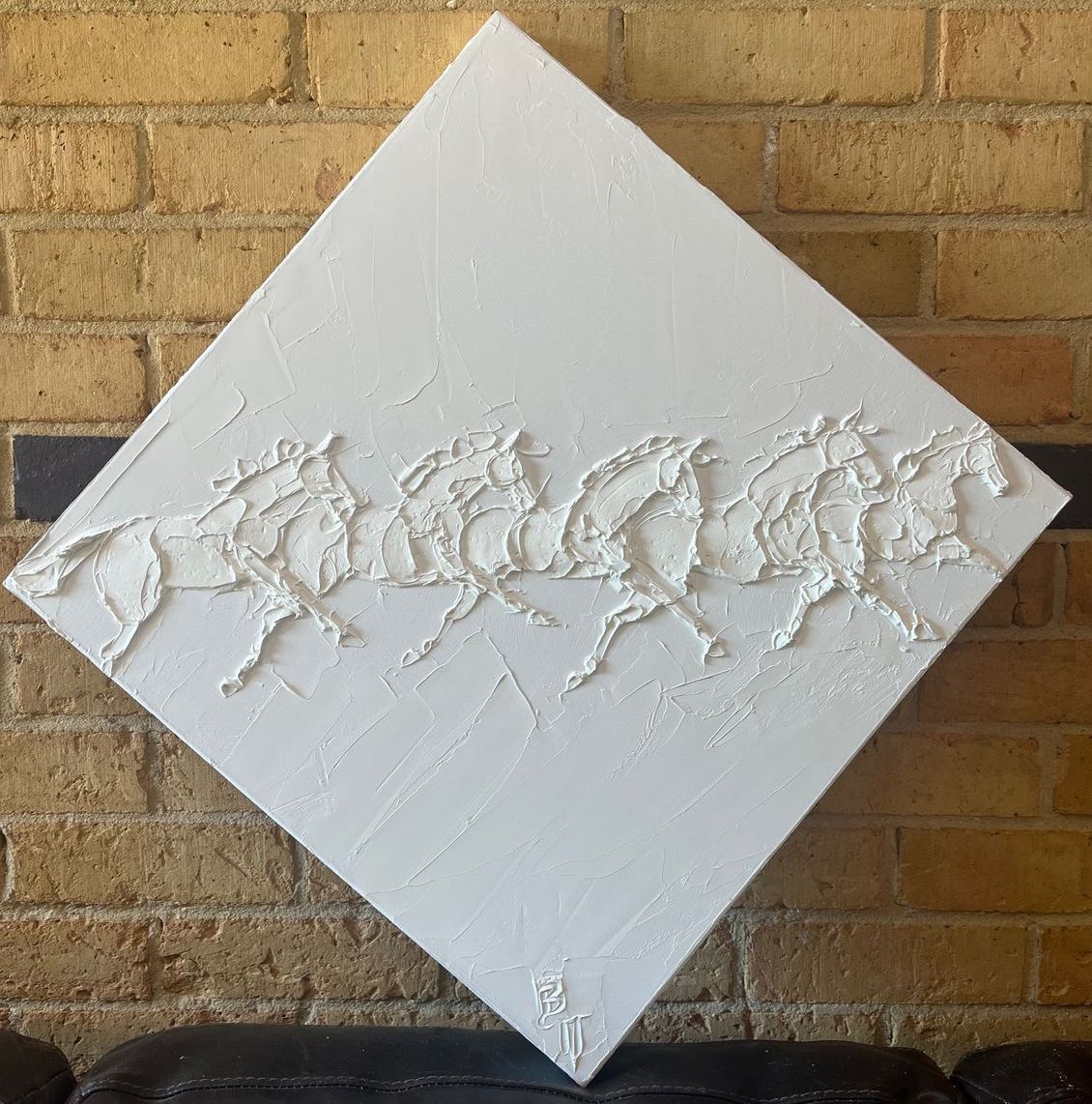 White textured painting of galloping horses on a diamond-shaped canvas, against a brick wall.