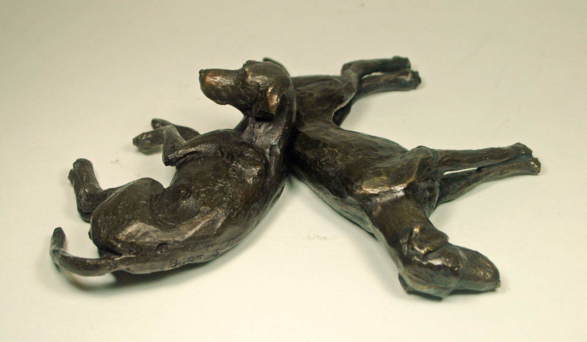 Two small bronze dog figurines lying on a light surface, touching nose to tail.