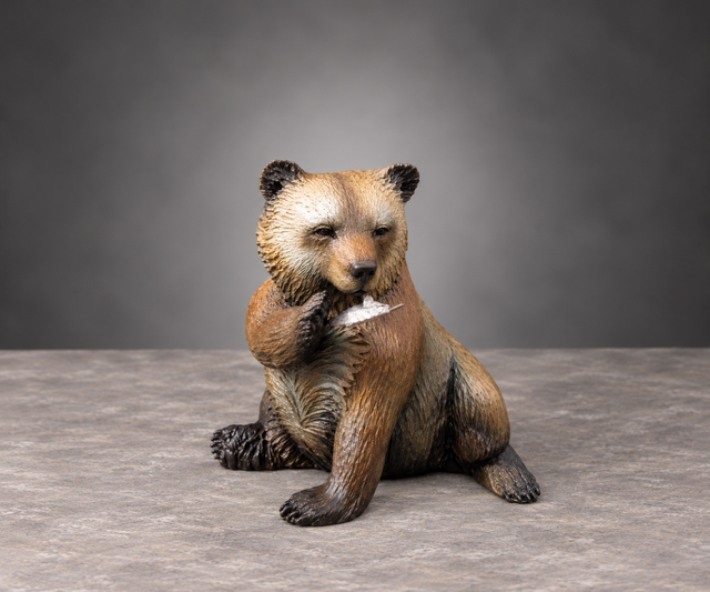 A small brown bear cub sitting on a gray floor against a gray backdrop, holding a white object in its paw.