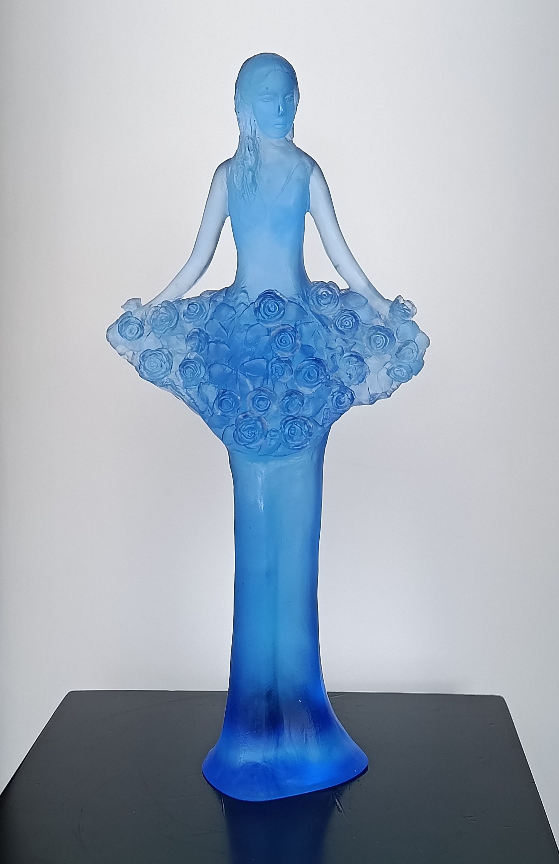 A blue glass sculpture of a figure with a textured, floral-style skirt, set against a plain light background.
