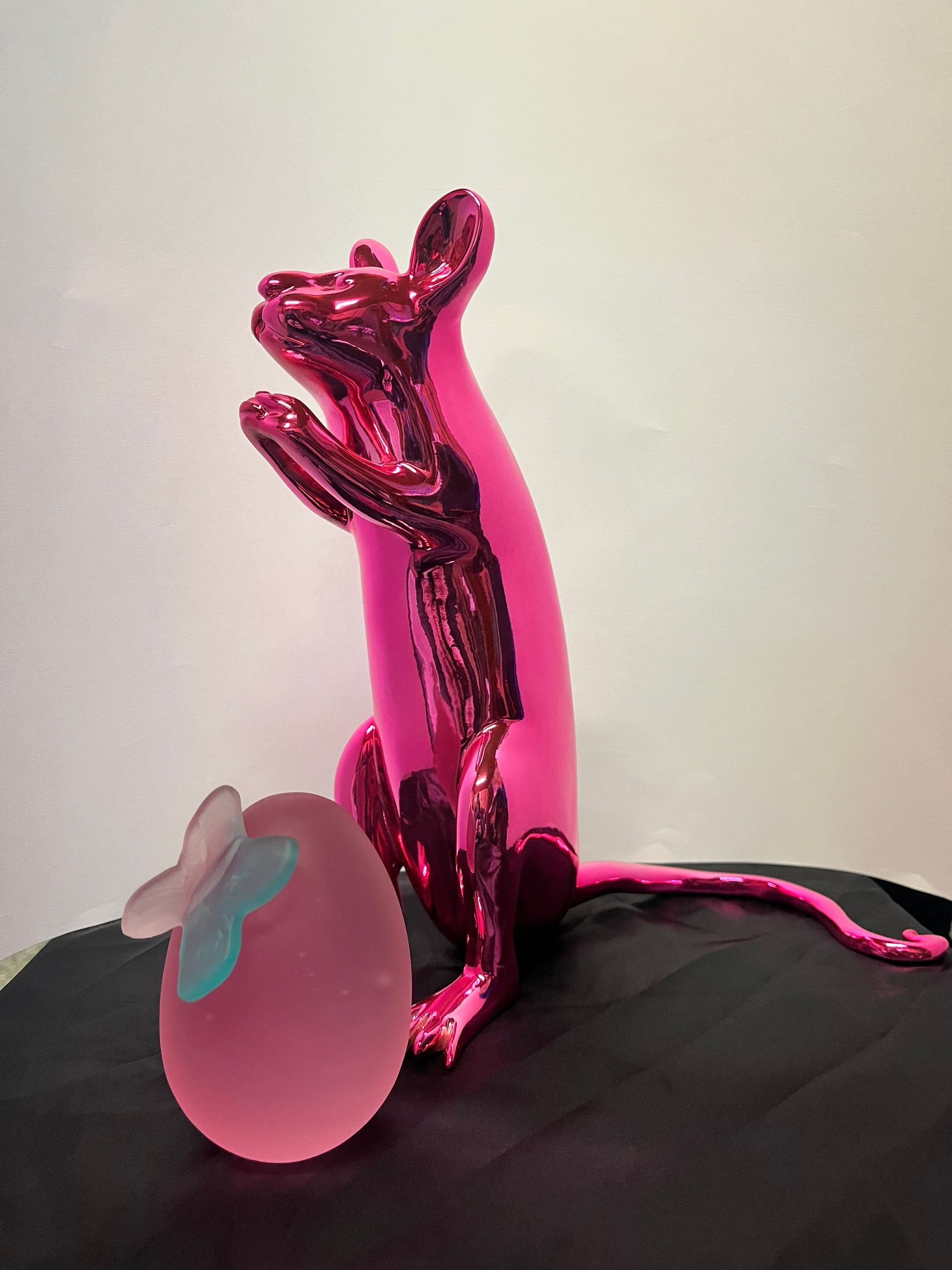 A shiny, bright pink sculpture of a standing mouse next to a translucent pink egg topped with a small blue butterfly.