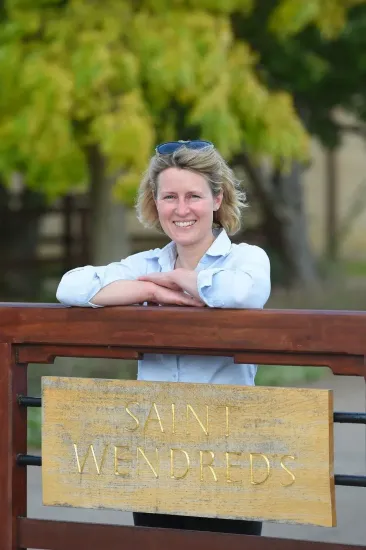 Woman leaning on gate with 'Saint Wendred's' sign; smiling, outdoors.