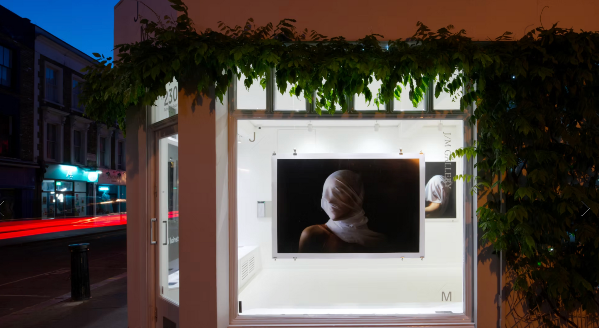 Outdoor art display in a lit booth at dusk, showing a portrait on a screen framed by hanging greenery