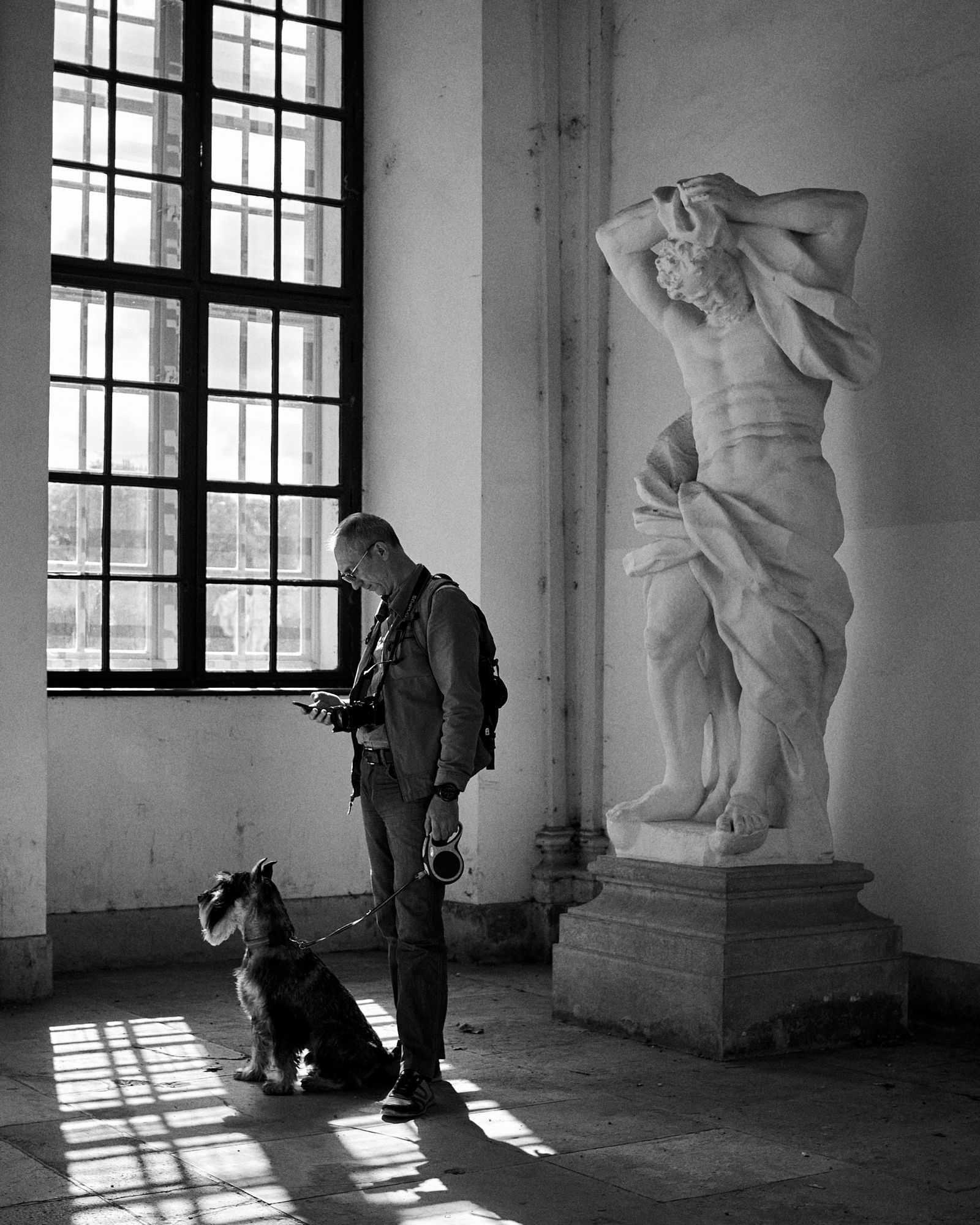 Man and dog beside a statue near a window, in a white-walled room, black and white.