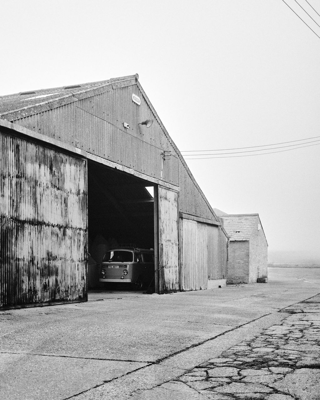 Black and white shot of a barn with open doors; a car is inside, with a cracked, concrete road.
