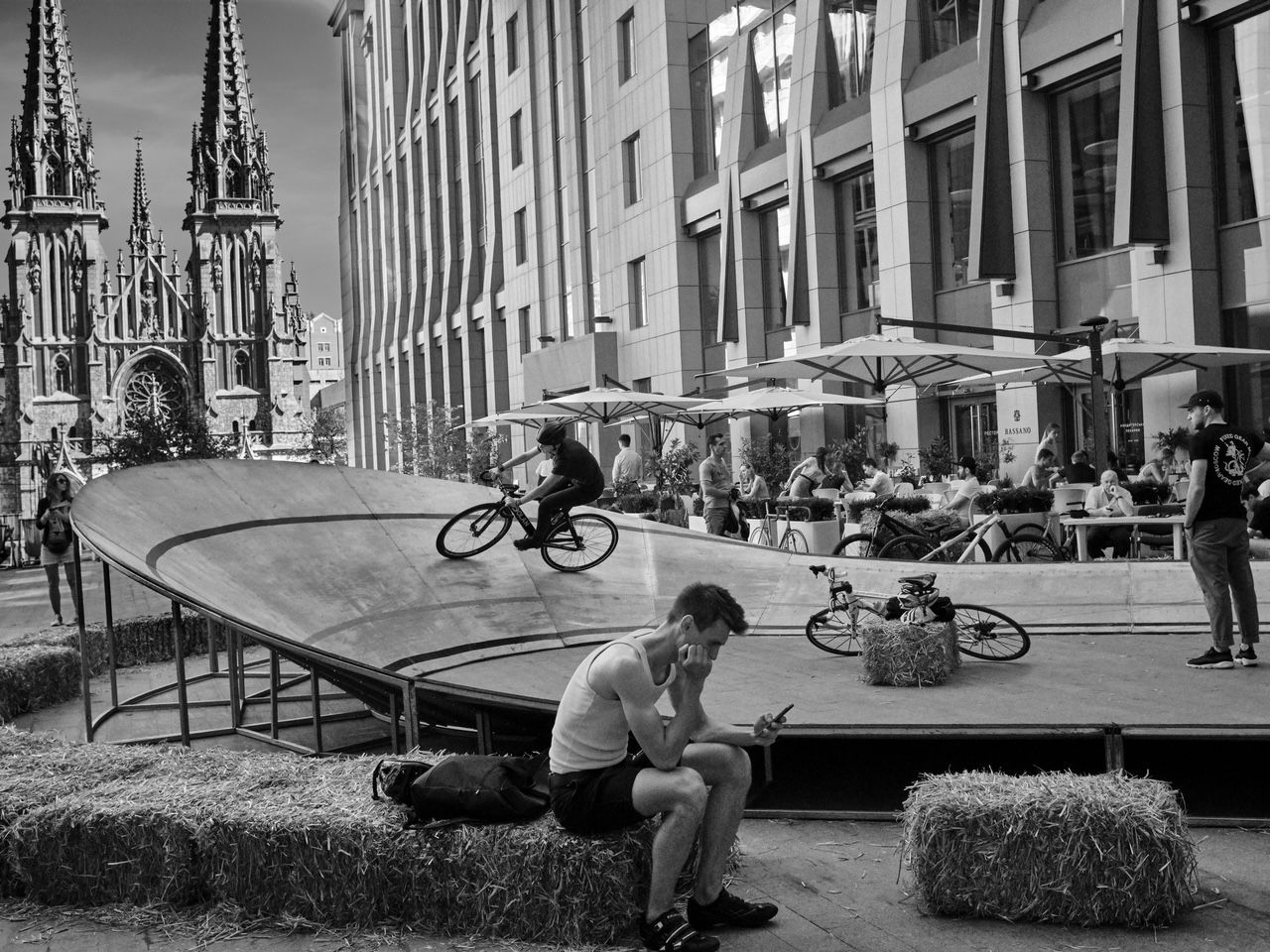 A biker on a ramp, people at tables, and a man using a phone. City setting with a cathedral in the background.