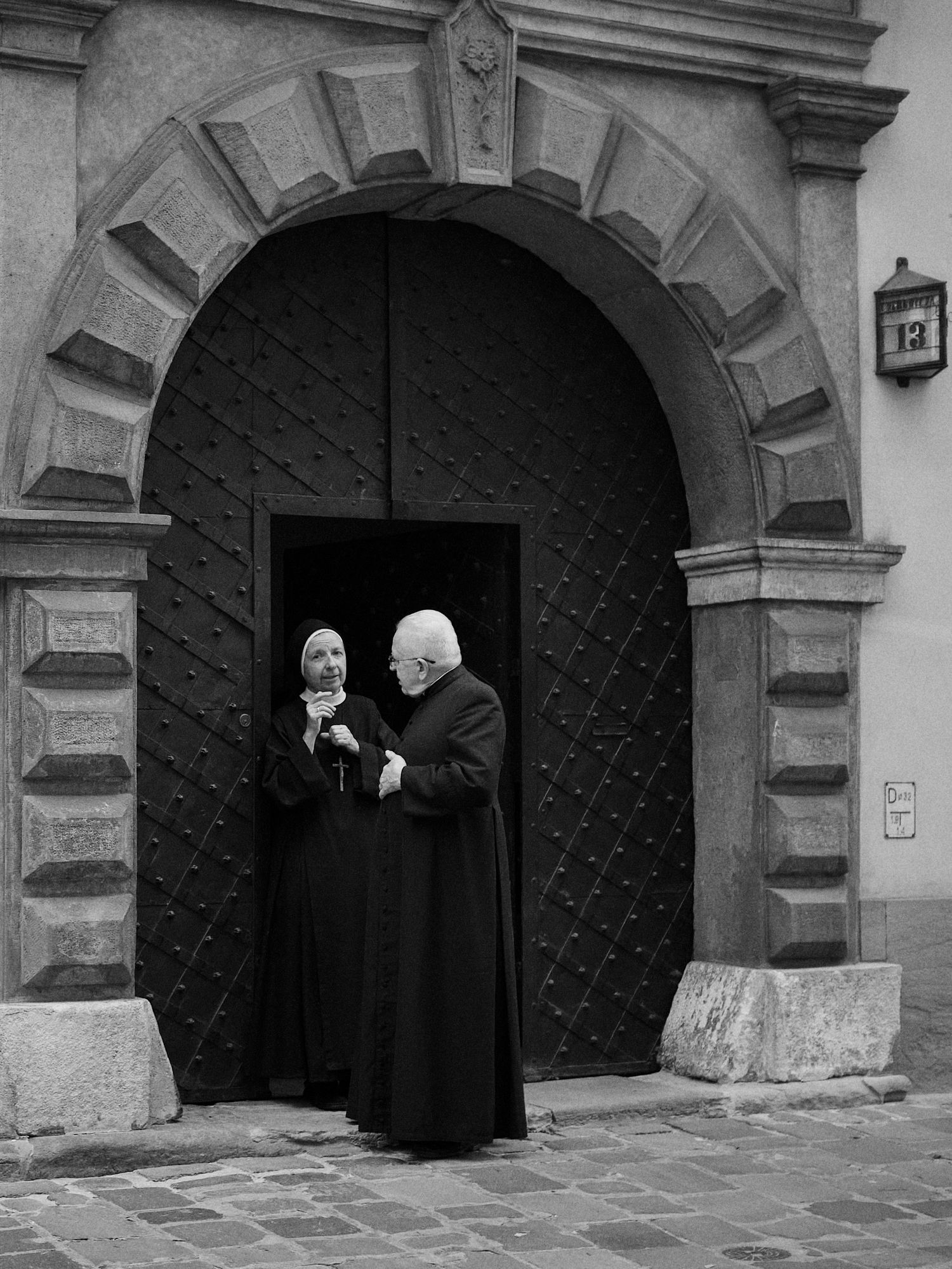 Two people, a nun and a priest, stand in a doorway with a large, arched door. Black and white.