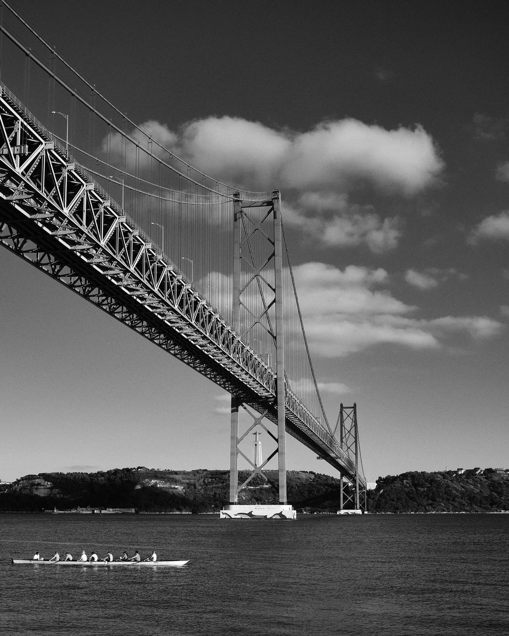 Black and white shot of a suspension bridge over water. A rowing team is on the water beneath the bridge.