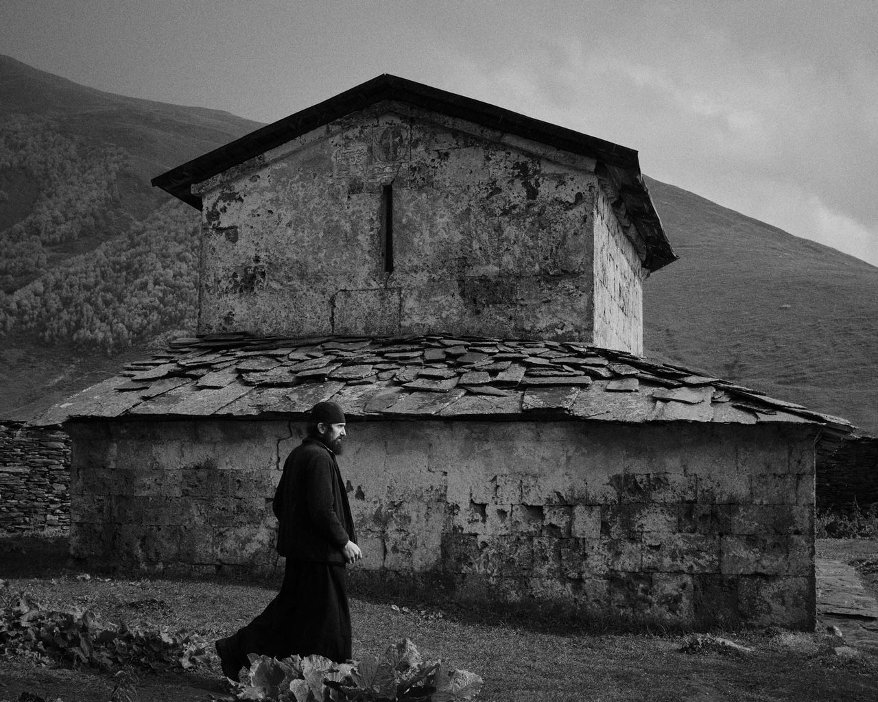 Monk in black robe walks past weathered stone church with slate roof, mountain backdrop.