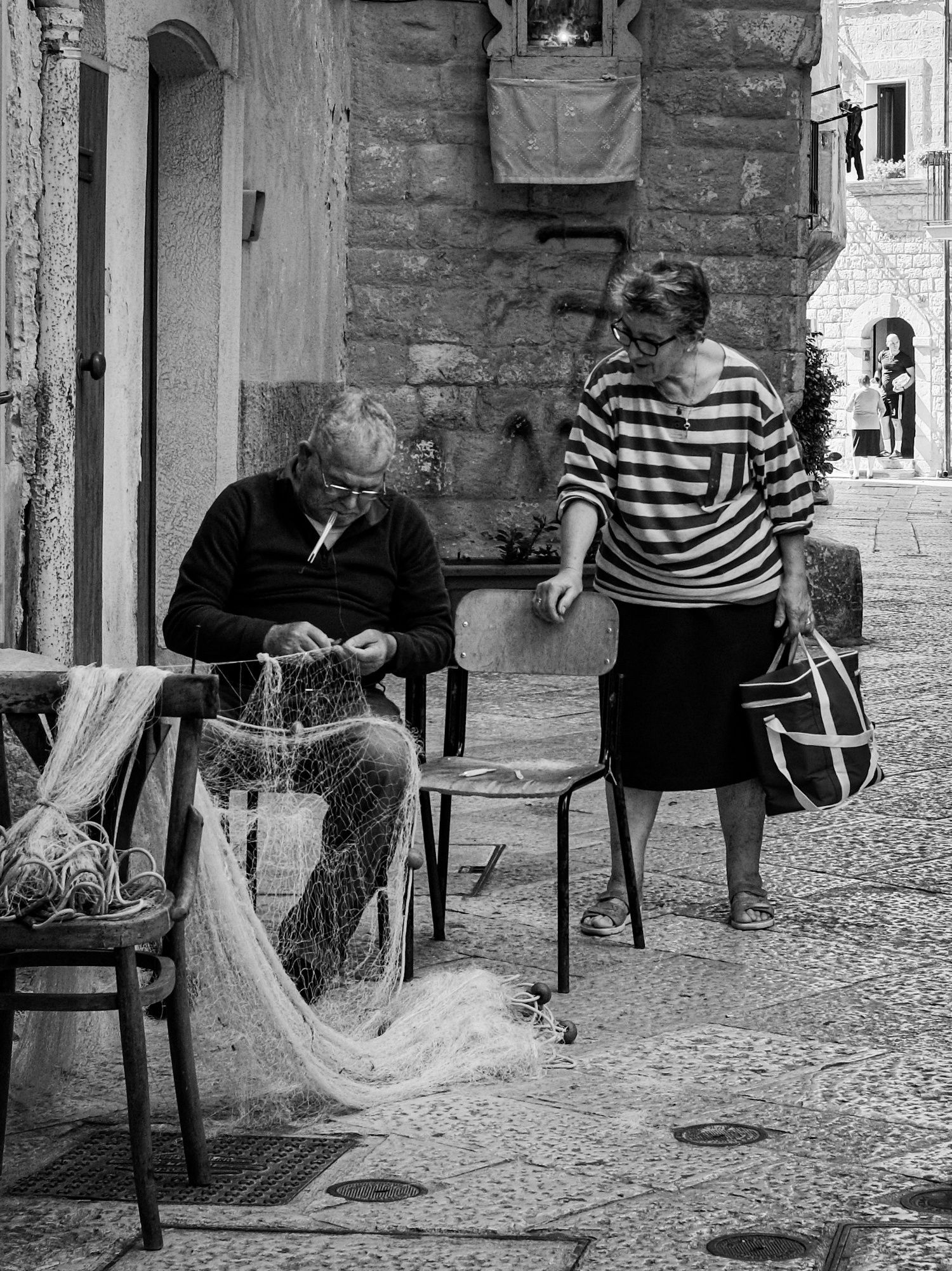 Elderly man mends fishing net in alley as woman looks on, holding bag. Black and white.