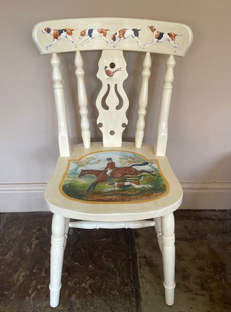 White painted chair with a hunting scene on the seat and dogs across the back, against a plain wall.