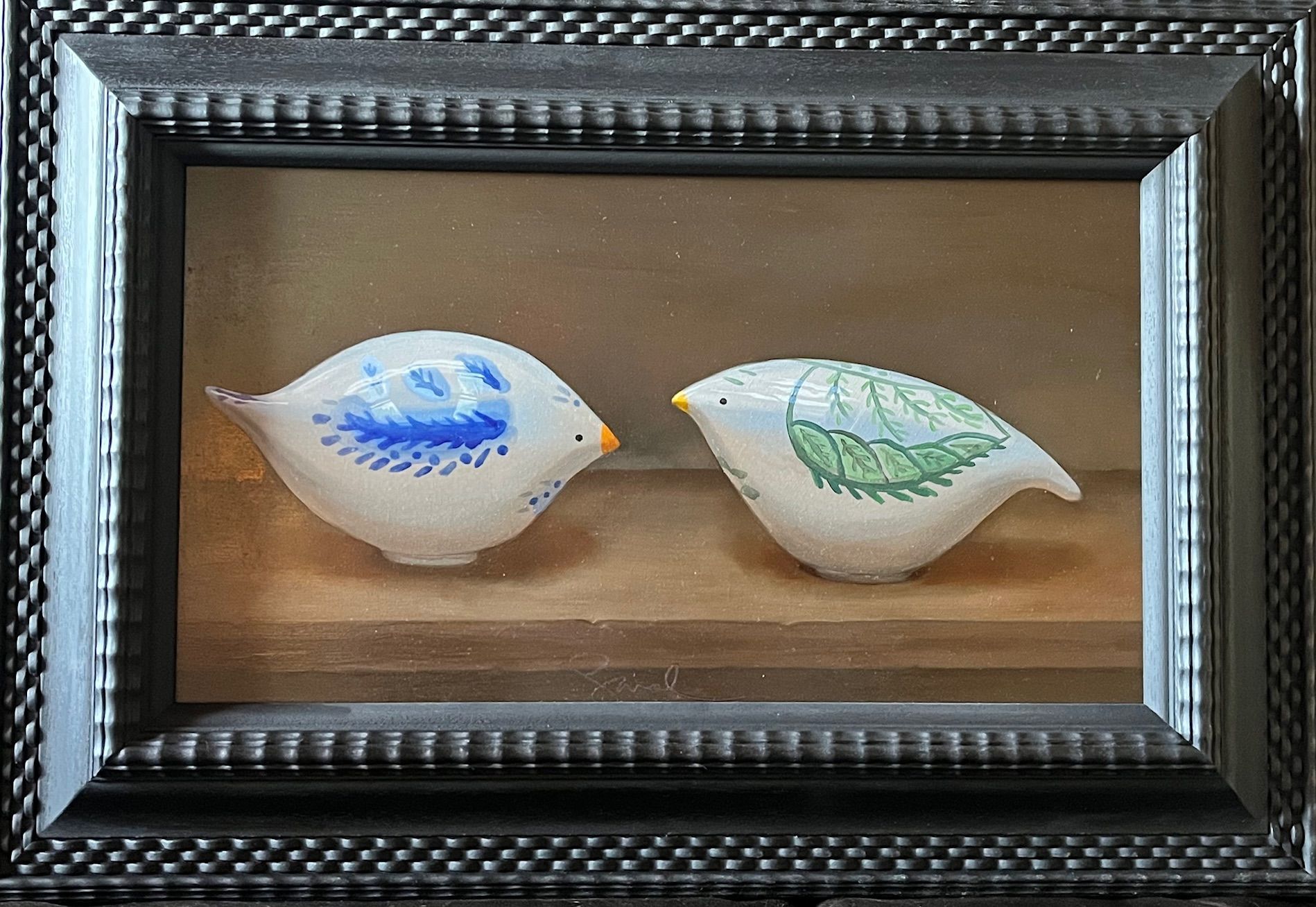 Two decorated white shell ornaments in a black frame, one blue and one green, on a brown background