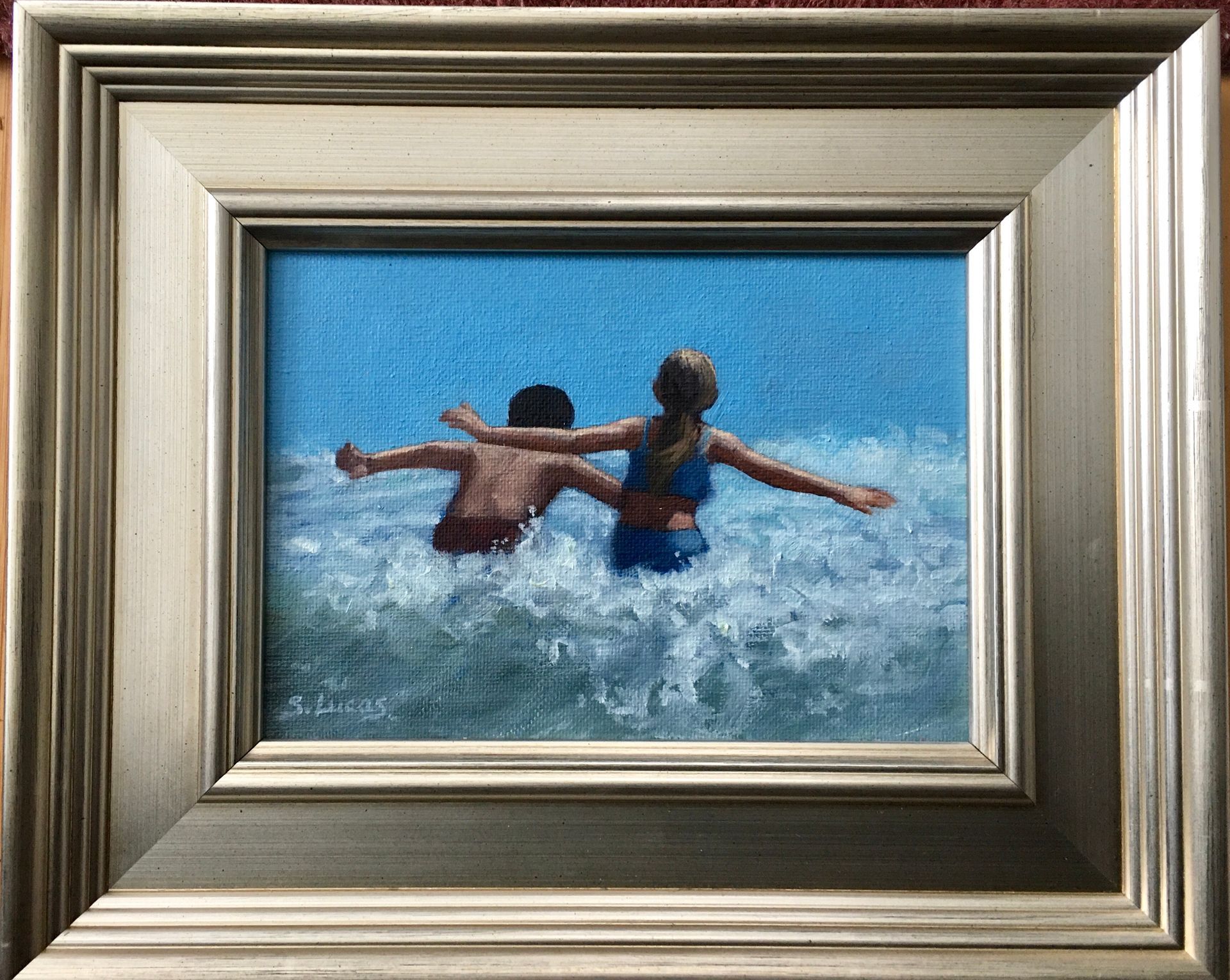 Two people with arms outstretched wade into blue ocean waves, depicted in an oil painting within a silver frame.