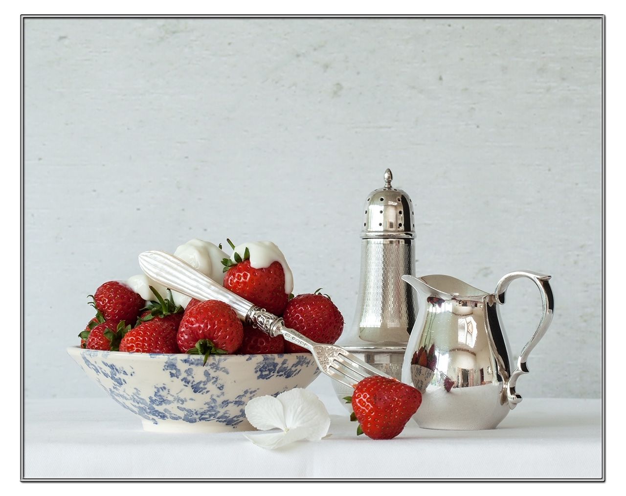 Bowl of strawberries and whipped cream, silver utensils and serving pieces on a white surface.