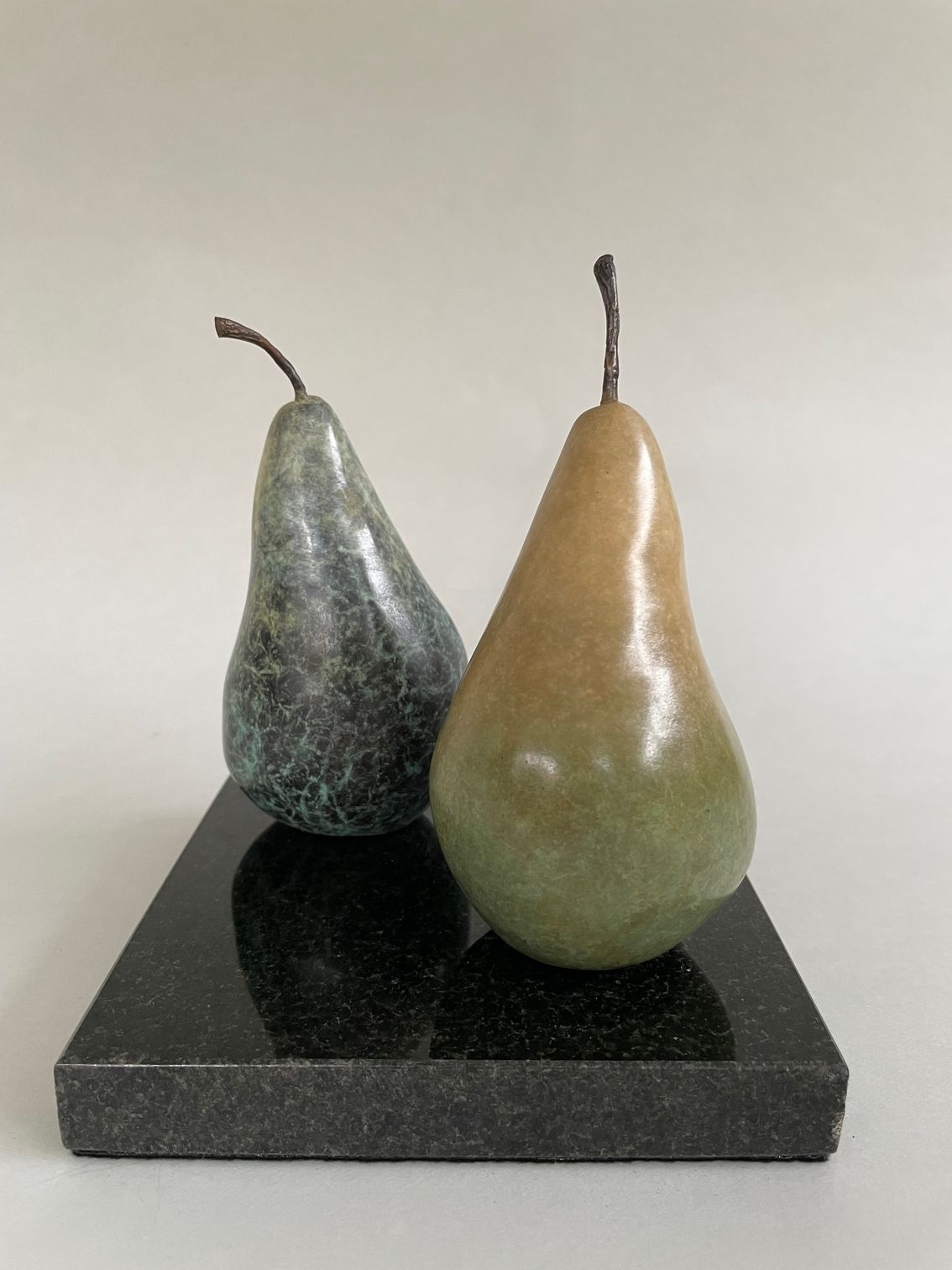 Two bronze pear sculptures, one dark green and one tan, displayed on a square black granite base against a light background.