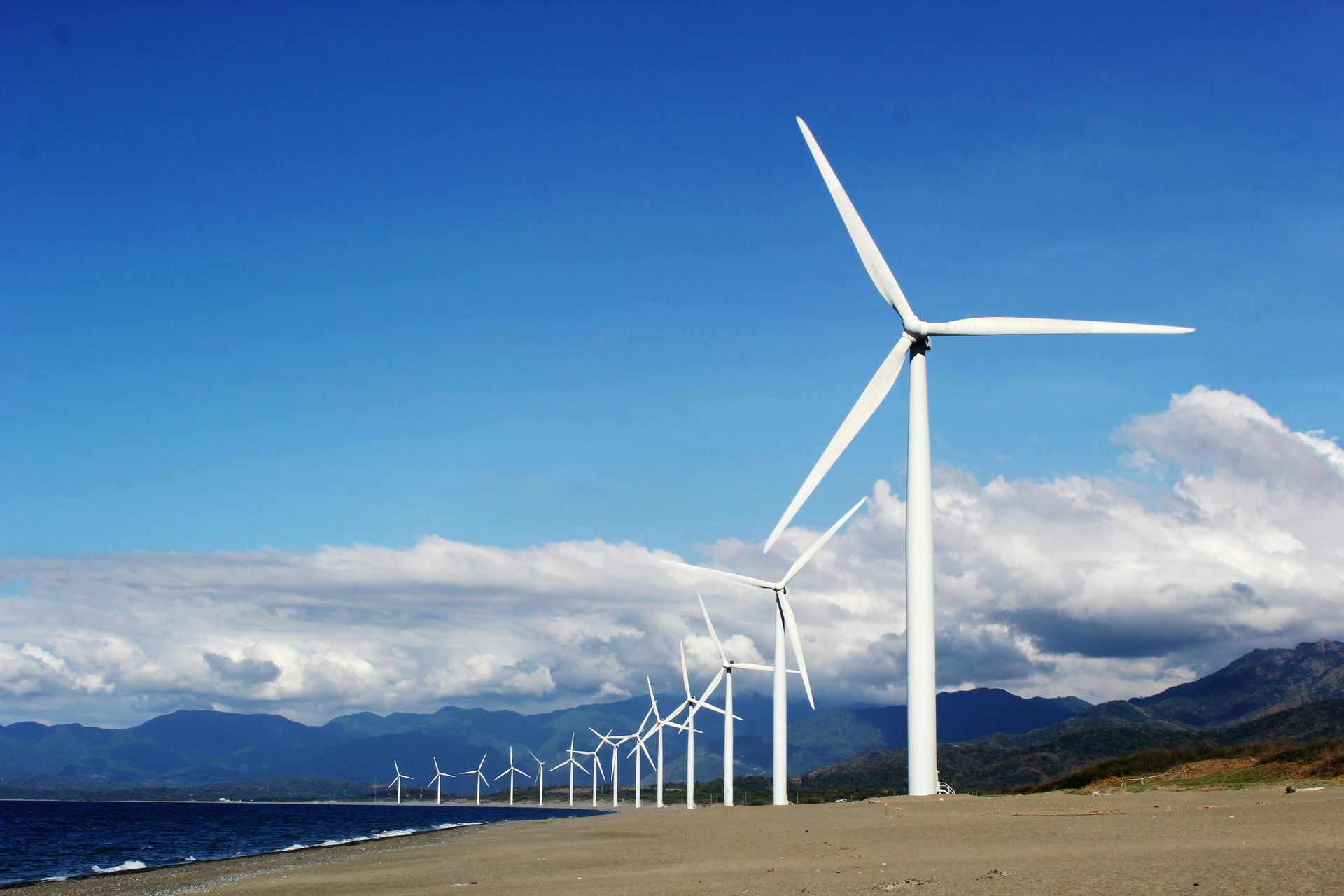 Wind turbines on a beach near mountains and ocean under a blue sky.