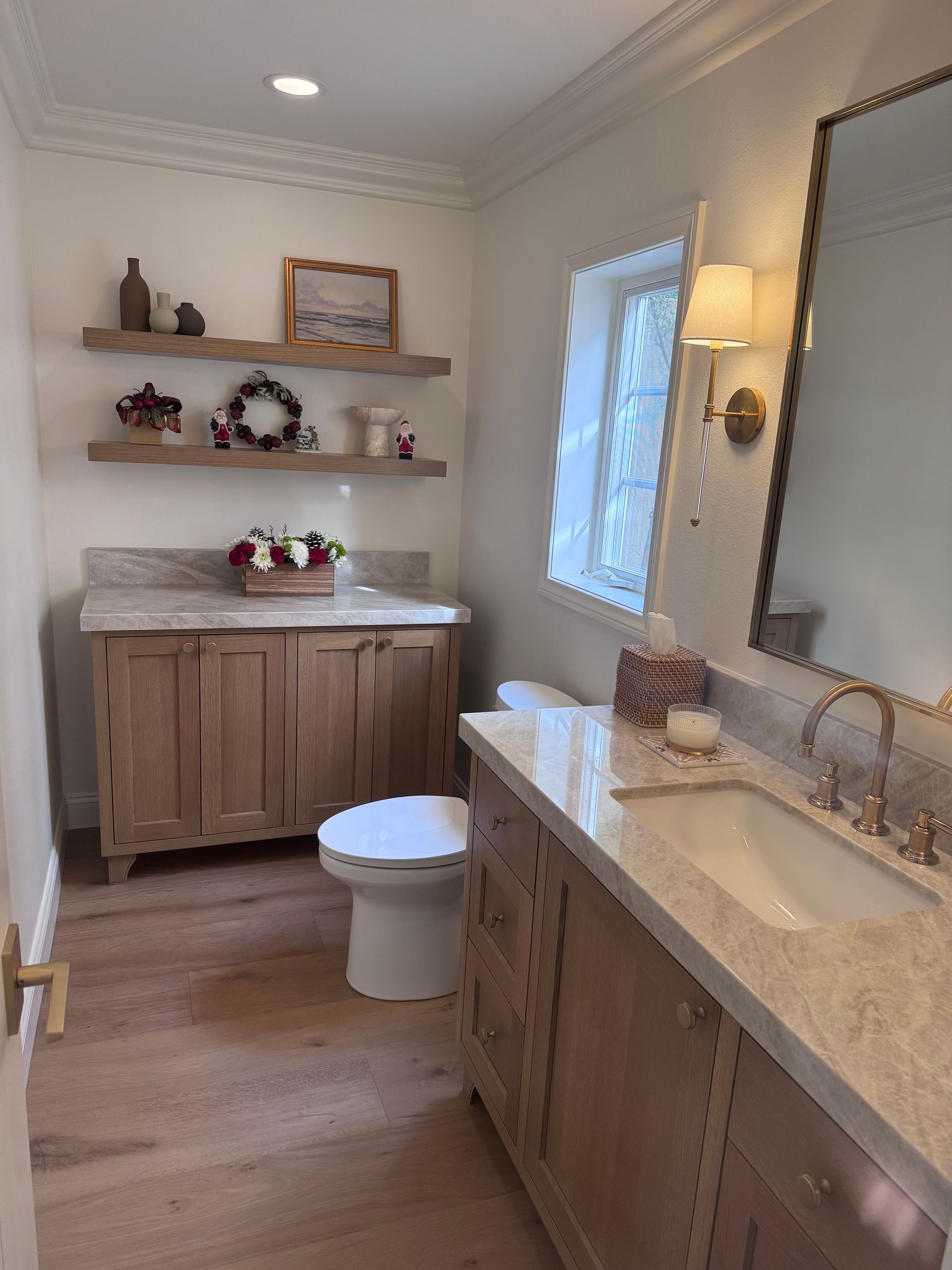 Bright bathroom with wood vanity, marble countertop, toilet, shelves, window, and large mirror.