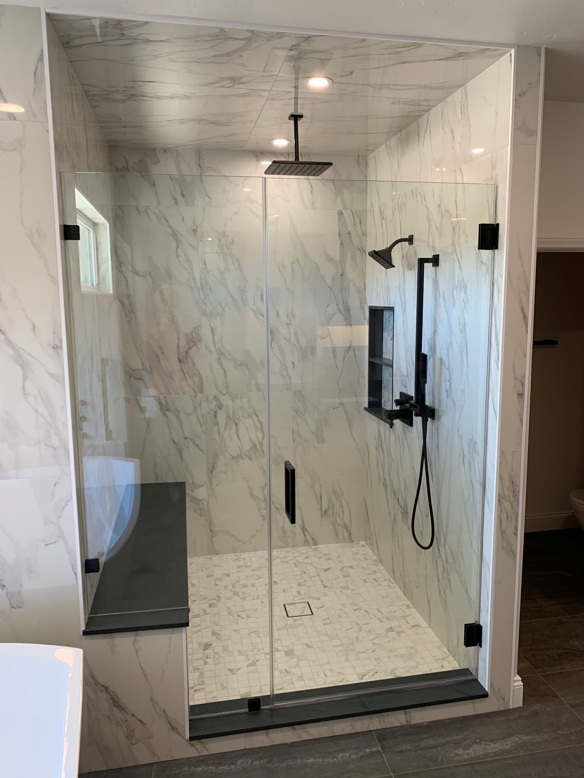 Modern marble-tiled shower stall with glass door, rainfall showerhead, handheld sprayer, and built-in bench.