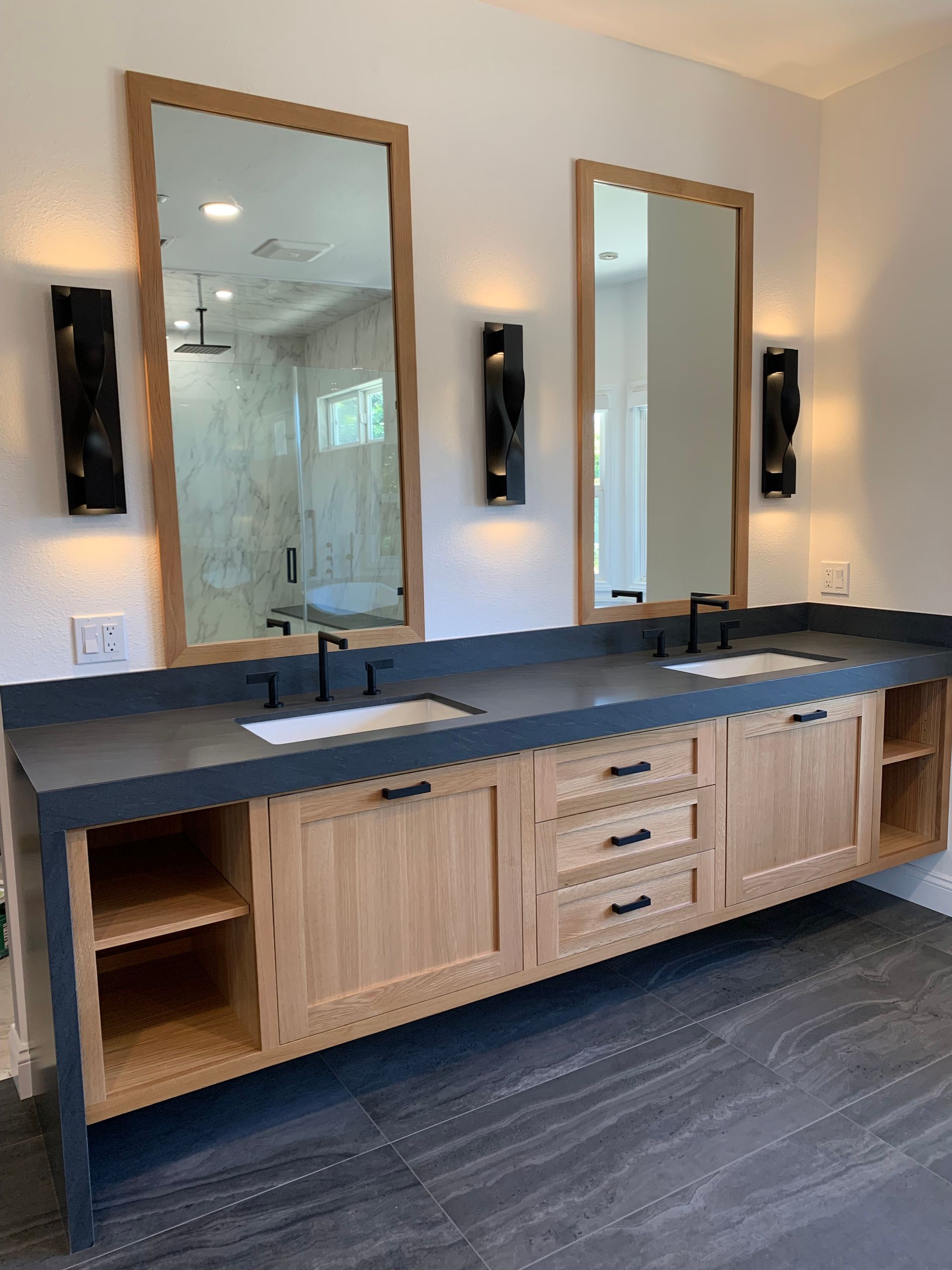 Modern bathroom vanity with two sinks, wood cabinetry, black countertops, and twin mirrors on a dark floor.