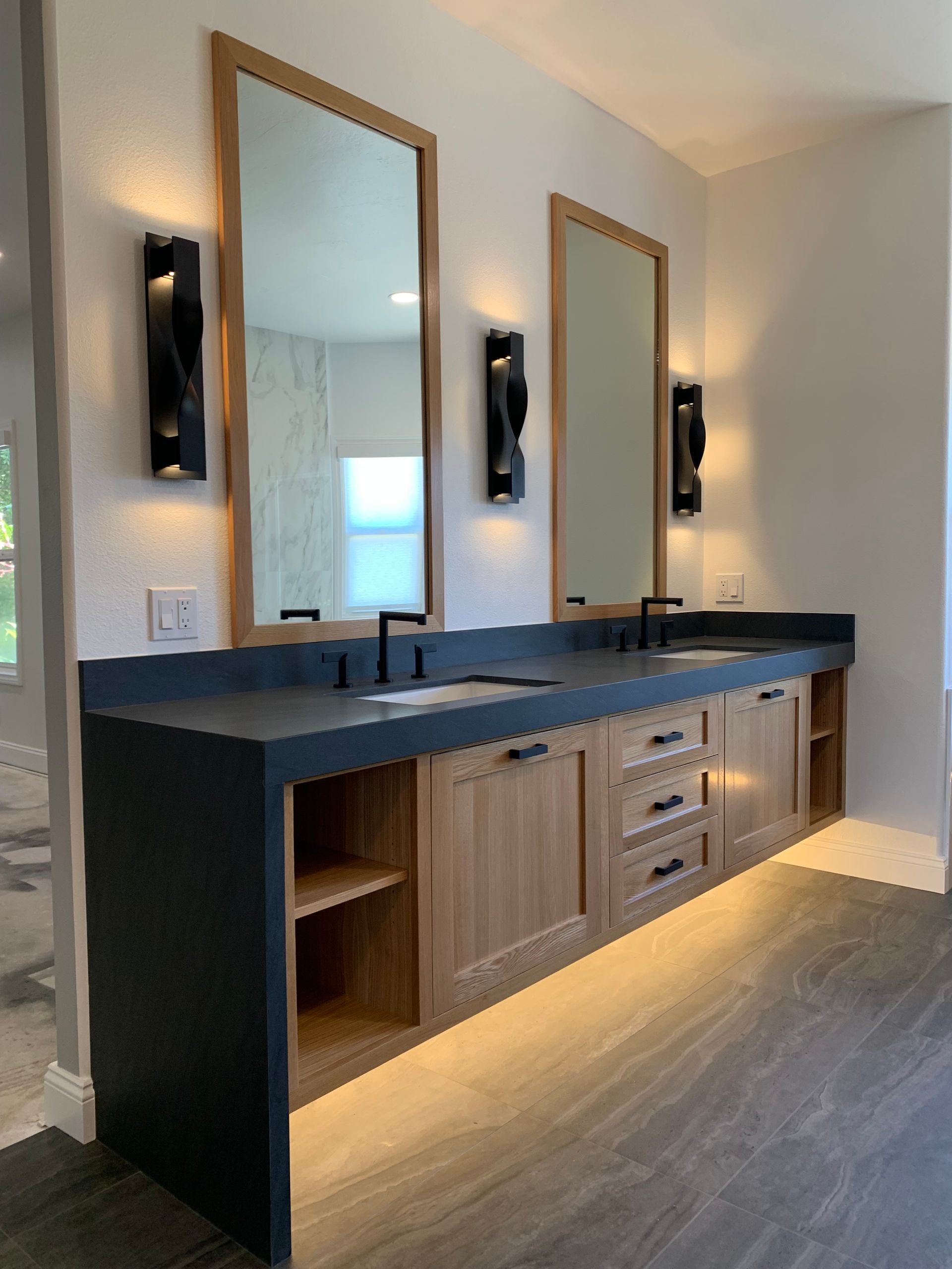 Modern double vanity with dark countertop, wood cabinets, and two mirrors in a bright bathroom.