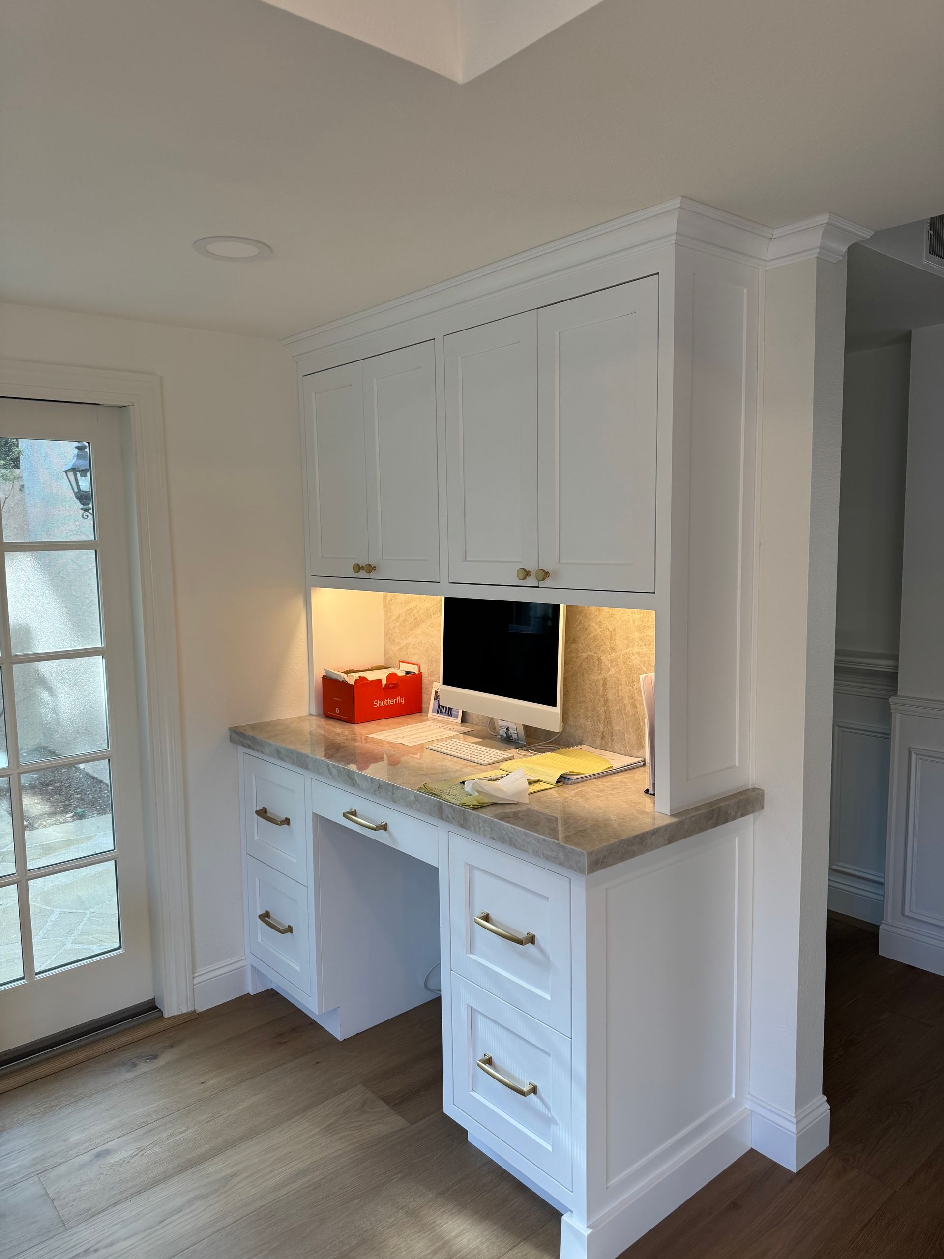 White built-in desk nook with cabinets, drawers, under-cabinet lighting, and a countertop in a bright room.