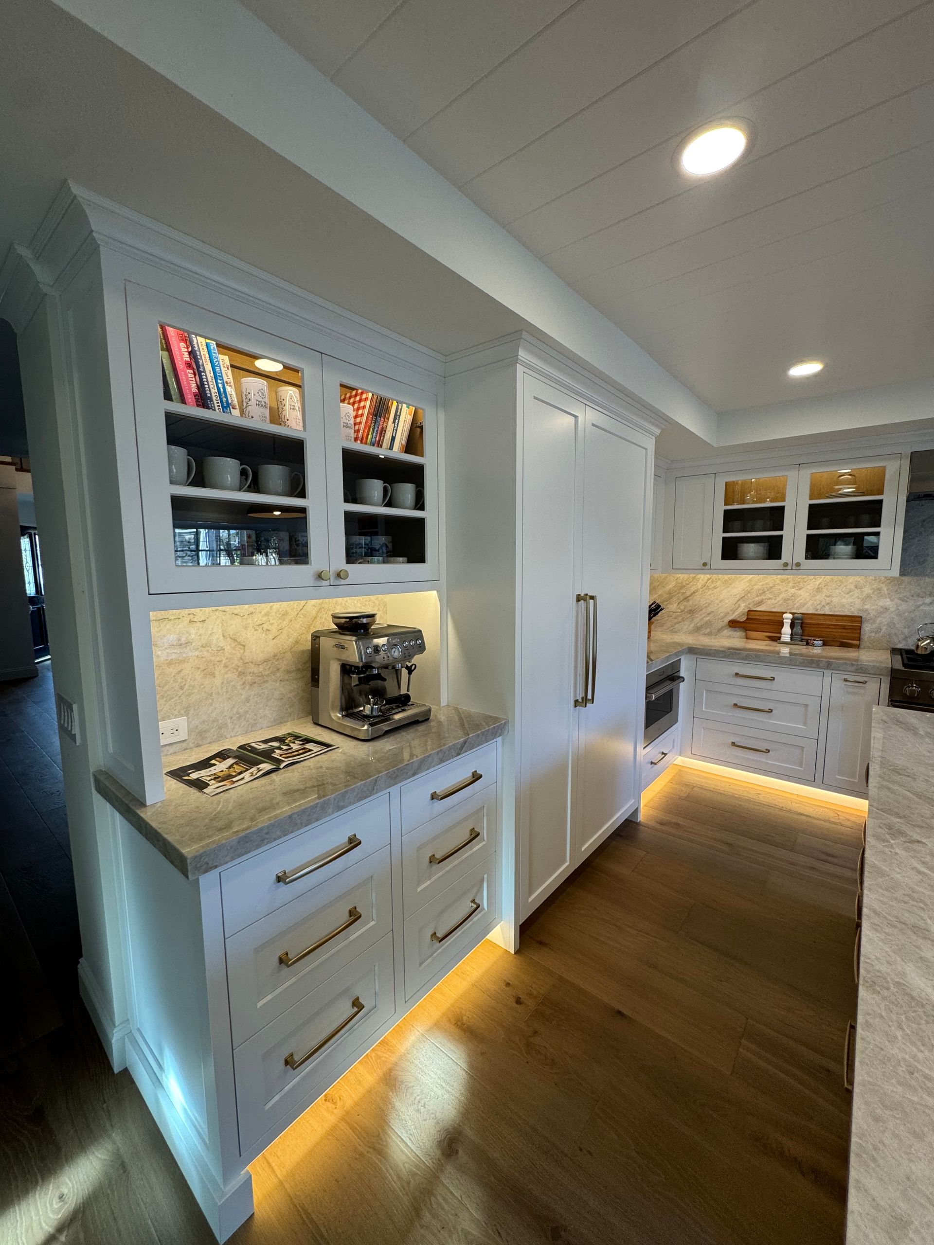 Bright white kitchen with built-in cabinets, coffee station, and hardwood floors lit by warm under-cabinet lights