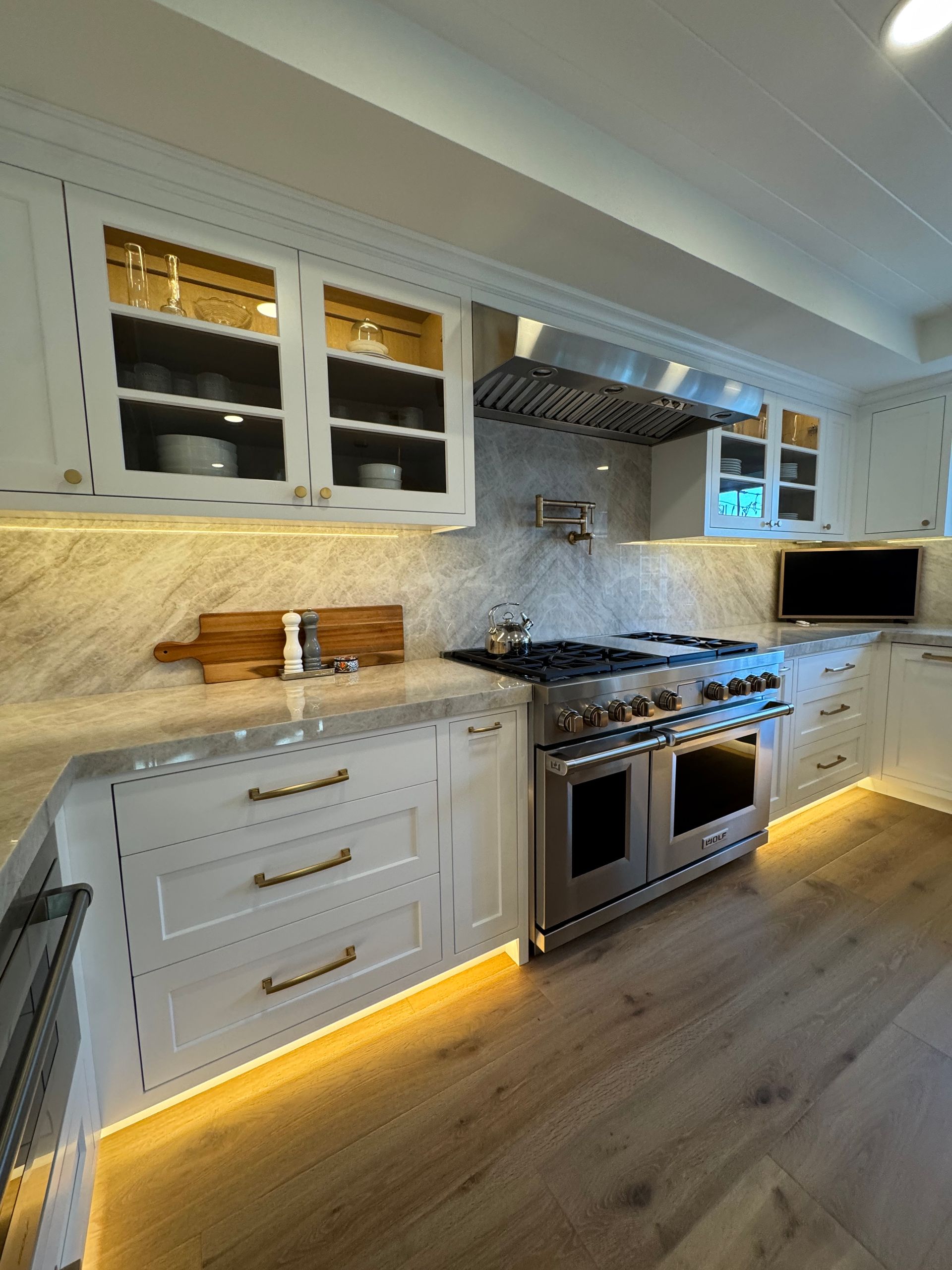Bright white kitchen with marble counters, stainless steel stove, lit under-cabinet lights, and hardwood floors