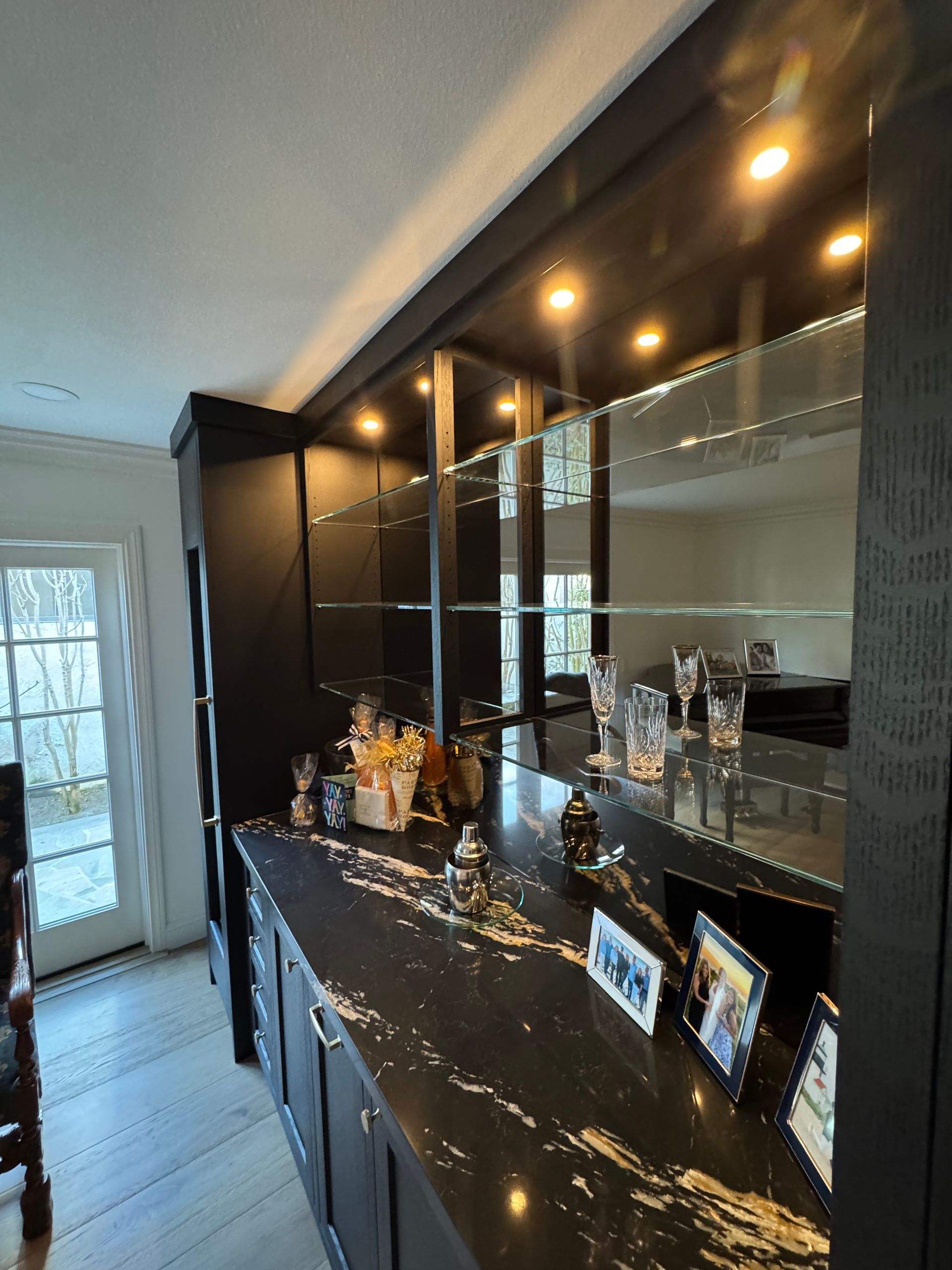 Glossy black bar cabinet with mirrored shelving, glassware, and framed photos in a bright hallway