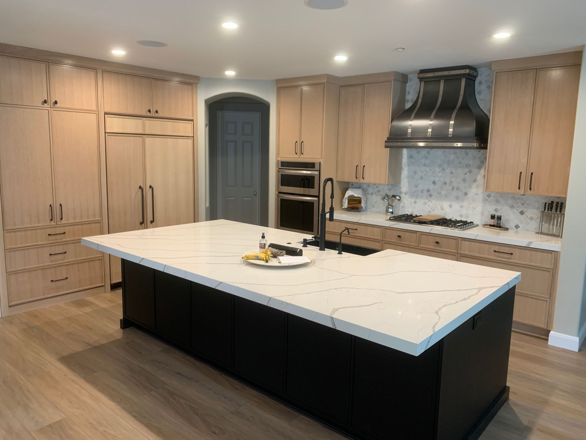 Modern kitchen with large white island, light wood cabinets, stainless hood, and built-in ovens