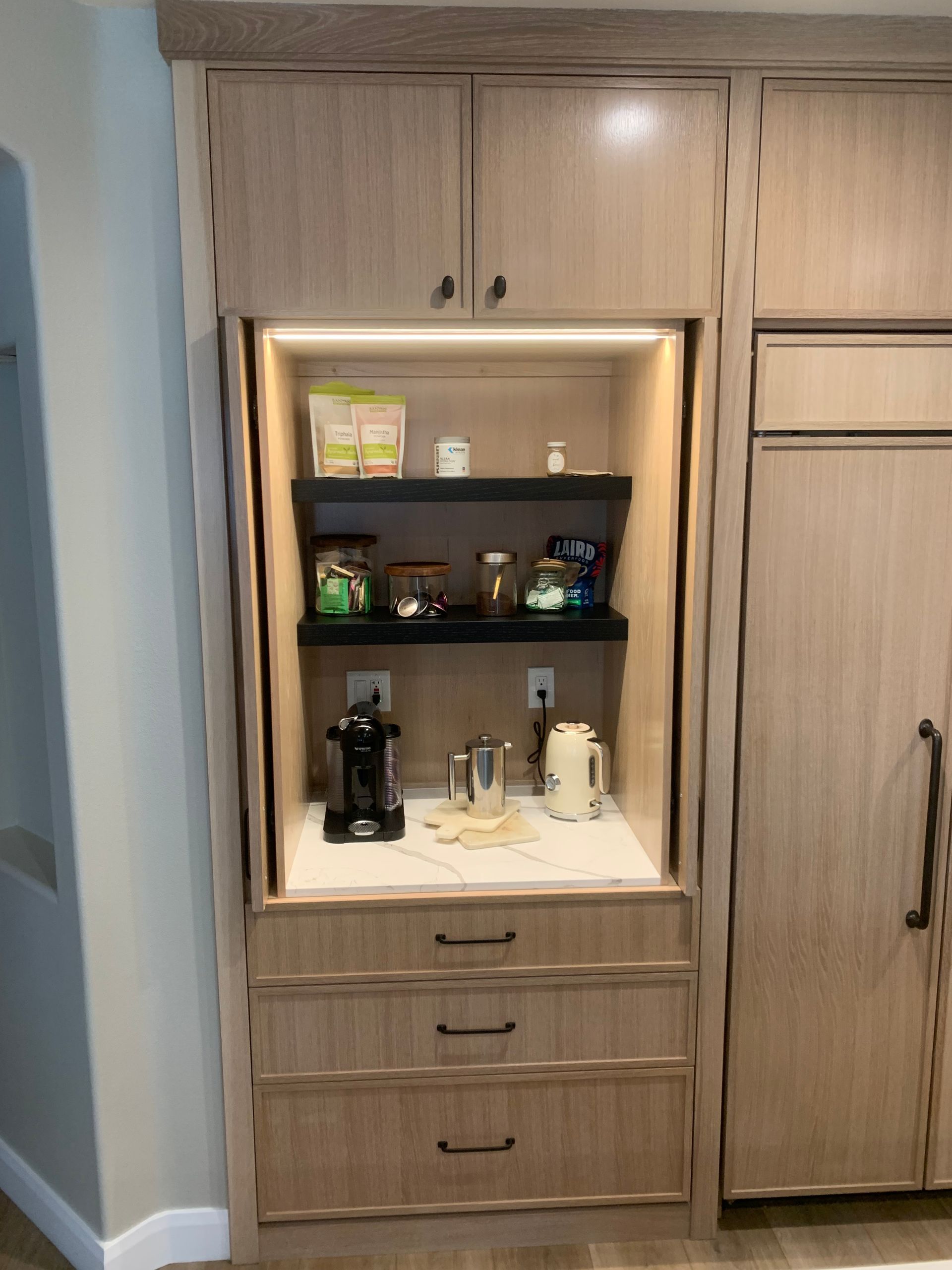Built-in kitchen coffee station with shelves, espresso machine, mugs, and drawers in a wood cabinet.