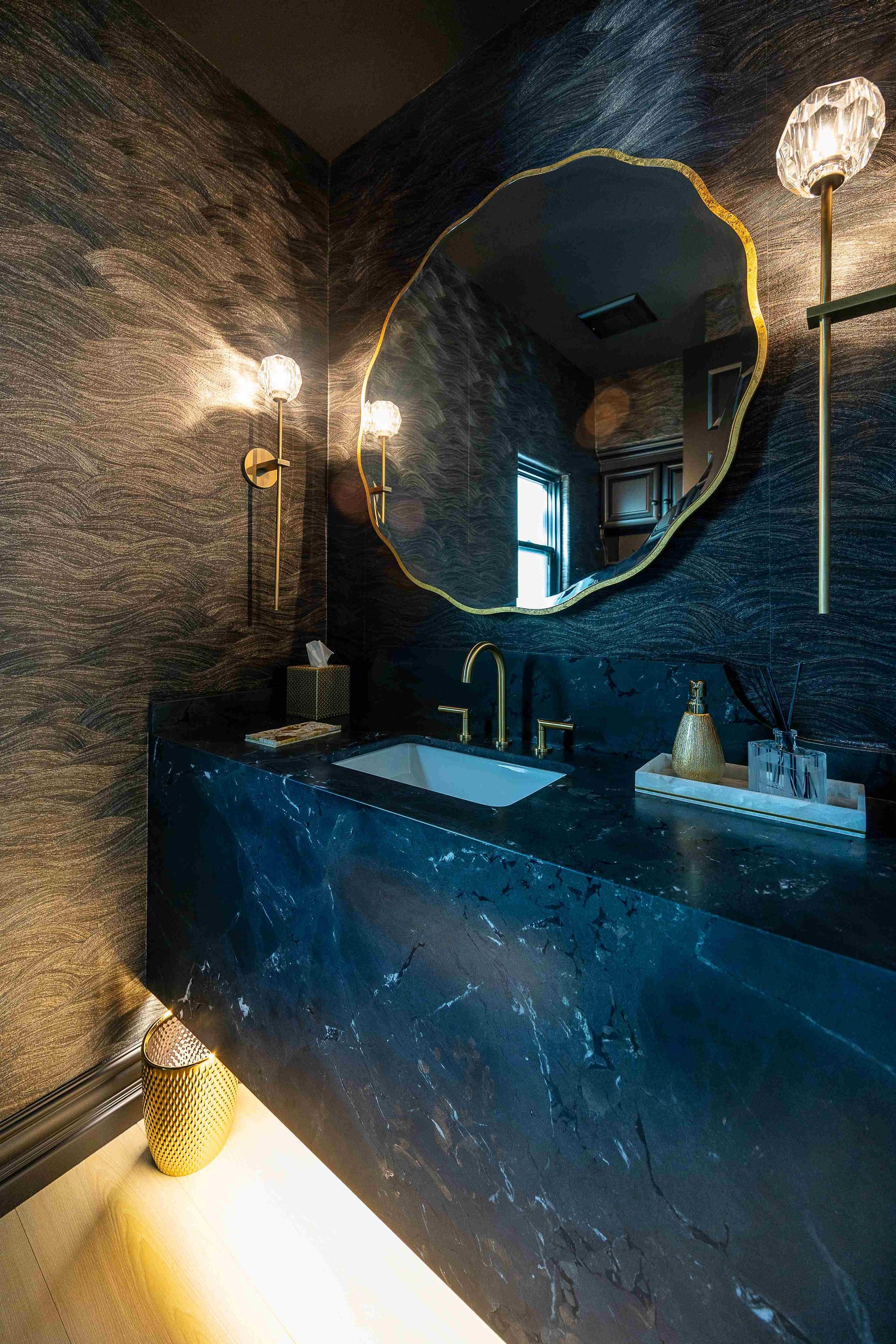 Dim luxury bathroom with textured dark walls, round mirror, glowing sconces, and a blue stone sink vanity.
