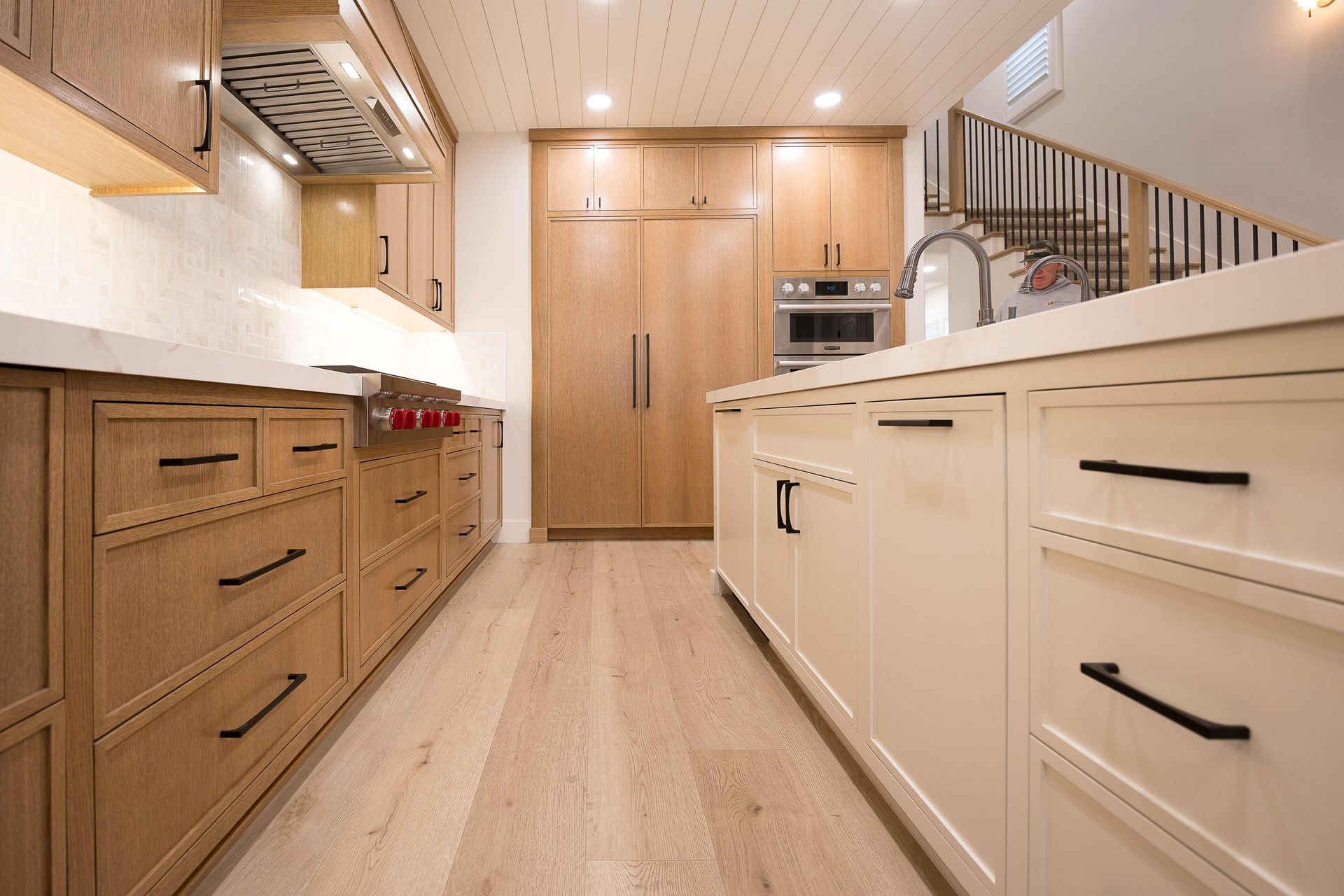 Modern kitchen with wood cabinets, white counters, and a light wood floor, viewed down a narrow aisle