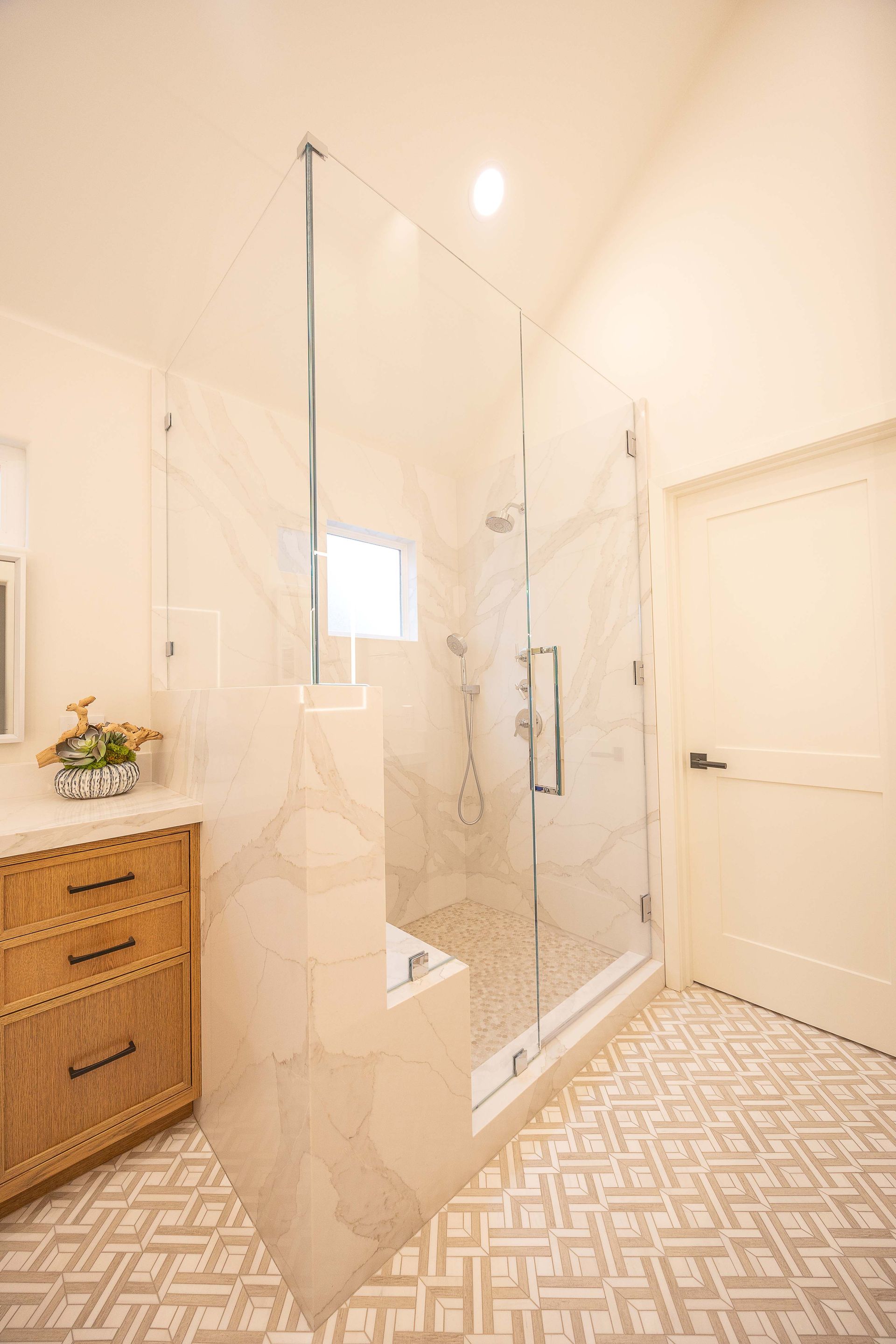 Bright bathroom with a glass walk-in shower, patterned tile floor, white walls, and a wicker cabinet.