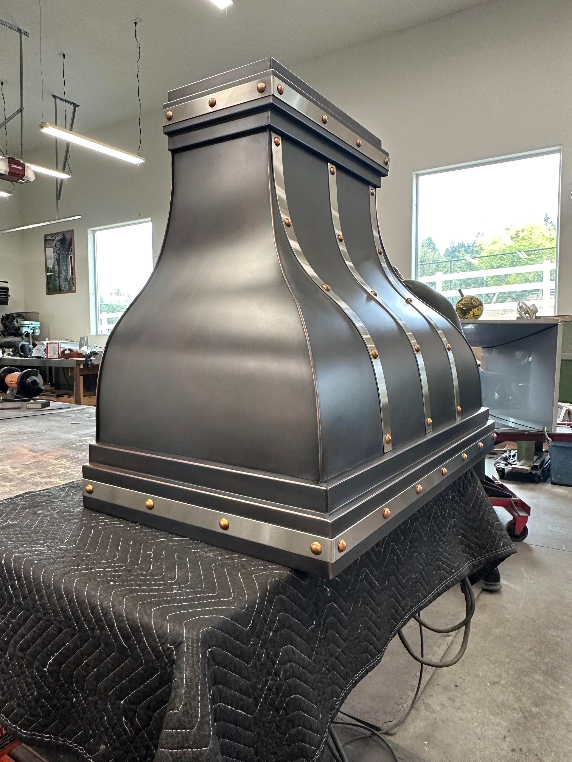 Large matte-black metal hood or chimney structure on a workshop table, with riveted edges and a windowed workshop background