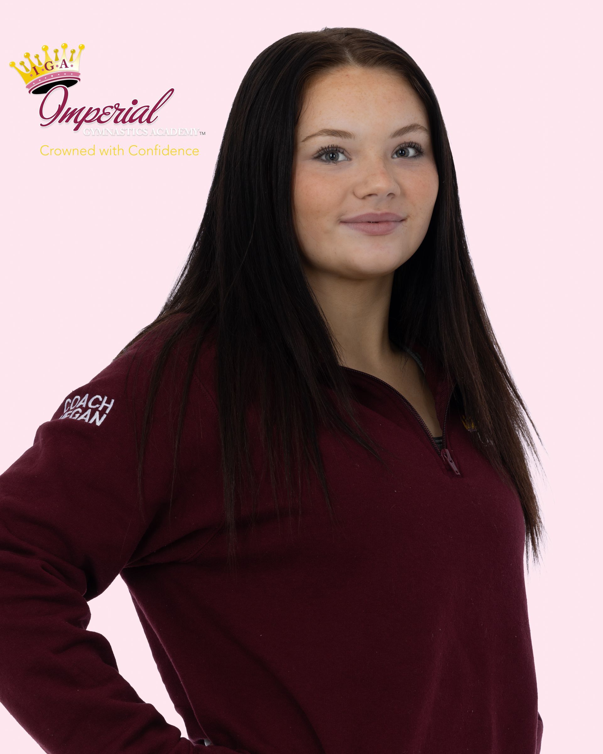 Woman with long black braids smiles, wearing a pink shirt with text, standing against a white wall.