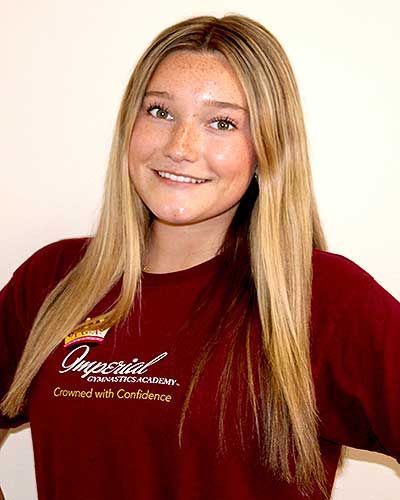 Woman with long dark hair smiles, wearing a pink t-shirt with text, necklace, against a white backdrop.