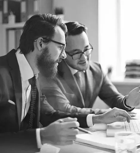 Two men in suits and ties are sitting at a table looking at a laptop.