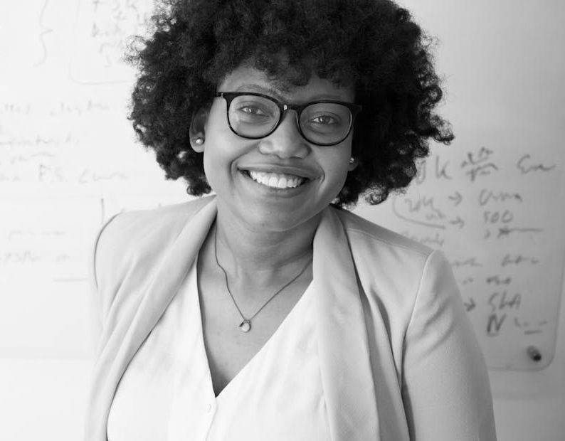 A woman wearing glasses and a white shirt is smiling in front of a whiteboard.